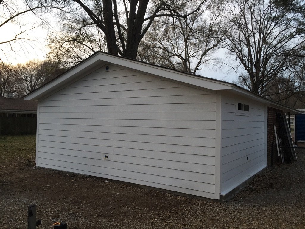 A small white detached garage or shed with horizontal siding, a sloped roof, and a small vent near the roof peak. There is a ladder leaning against the side, and the ground around is dirt.