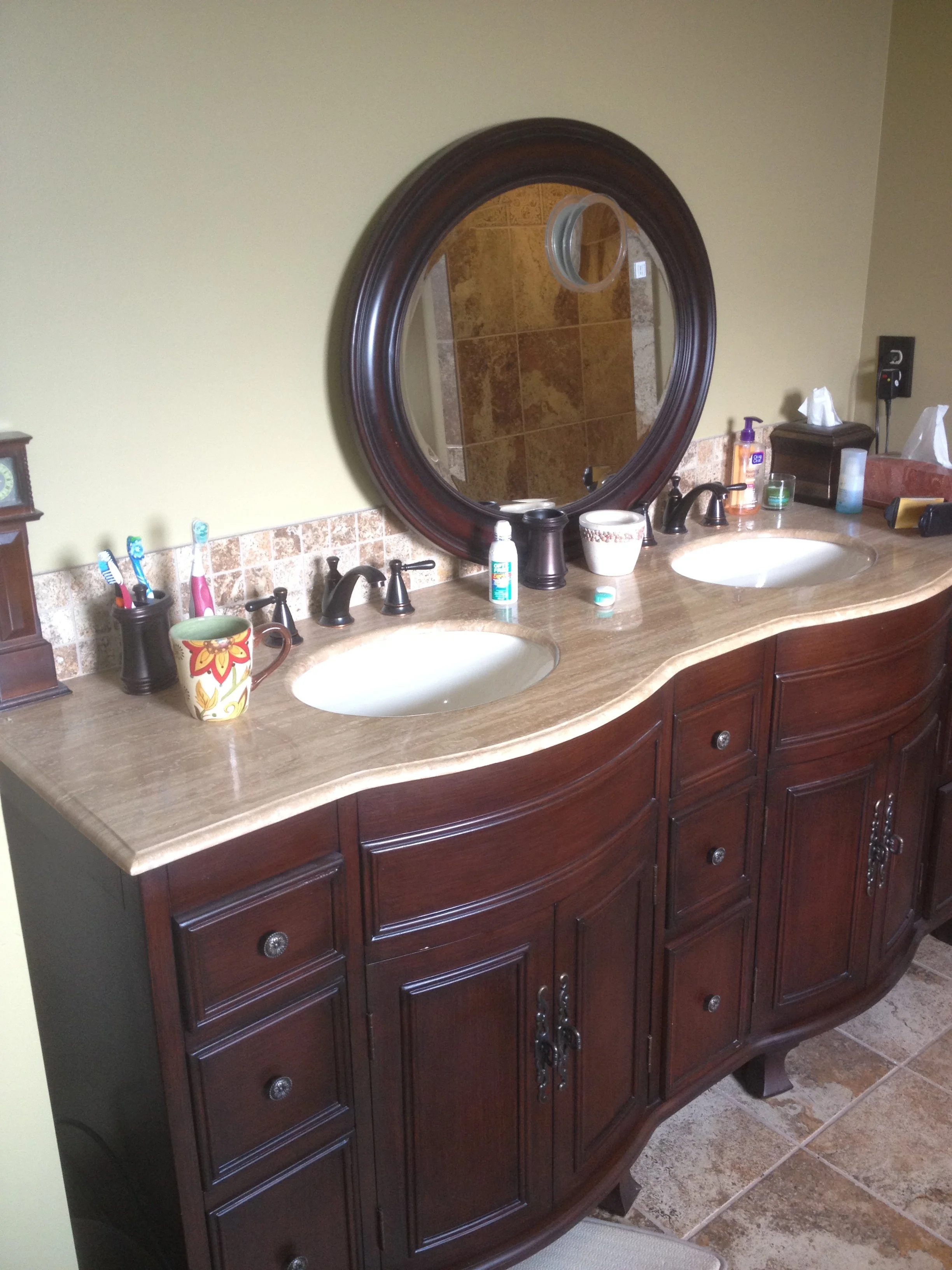A bathroom vanity with a marble top, two sinks, and oval mirror, with items like toothbrushes, hand soap, tissue box, and other toiletries on the counter.