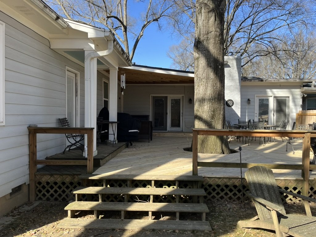 A backyard deck attached to a white house, with steps leading up to it, surrounded by trees with no leaves, and outdoor furniture including chairs and a table.