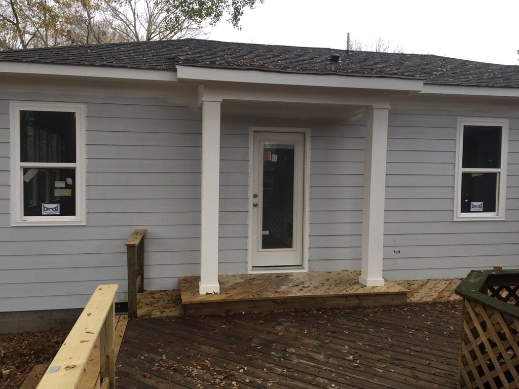 Backside of a house under construction with a newly installed small wooden deck, white exterior siding, double-hung windows, and a glass door leading to the deck. The deck has two support columns and a partial railing on the left side, with some leav