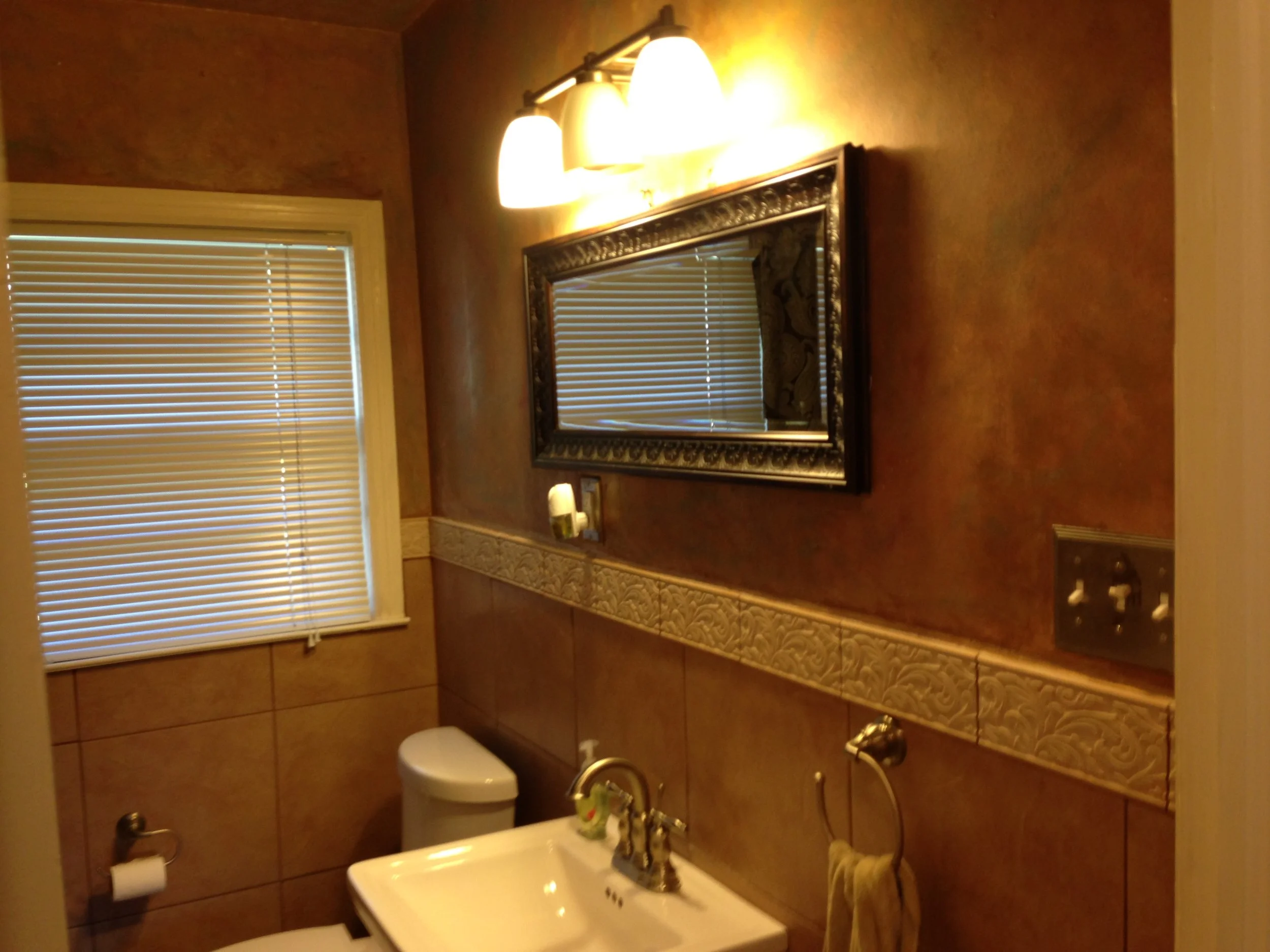 A bathroom with a window with closed blinds, a mirror mounted on a wall with a light fixture above it, a toilet, a small sink with a gold-colored faucet, and a decorative border tile. There is a towel hanging next to the sink and a soap dispenser on 