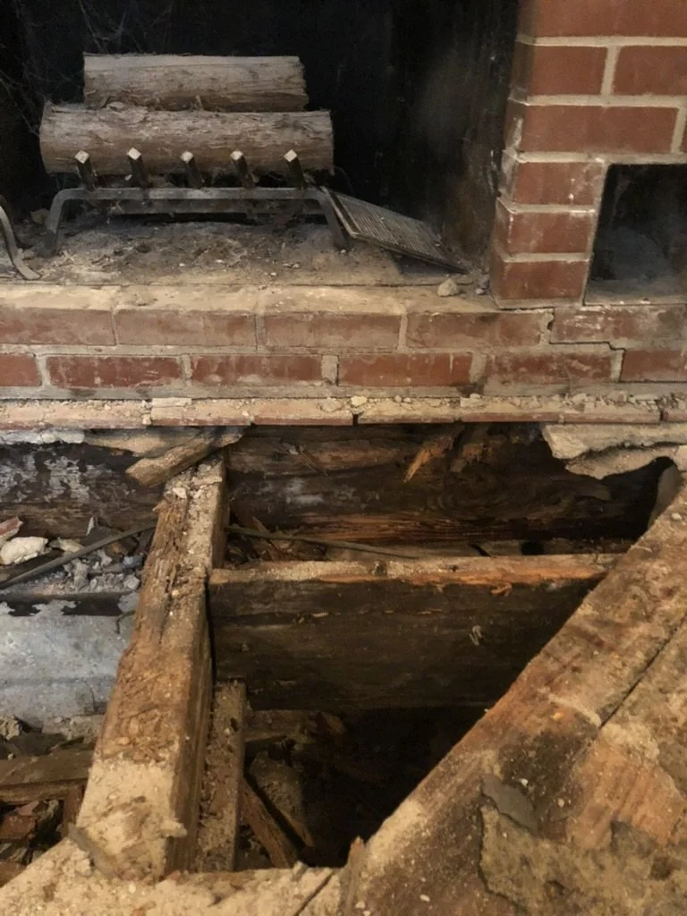 A fireplace with missing brick and exposed aged wooden support beams, some logs placed on a metal grate inside the fireplace