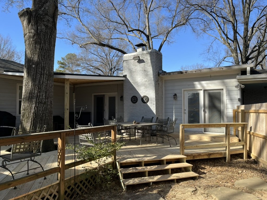 A backyard deck with patio furniture, a chimney, a large tree, and a sliding glass door leading into the house, surrounded by a wooden fence.