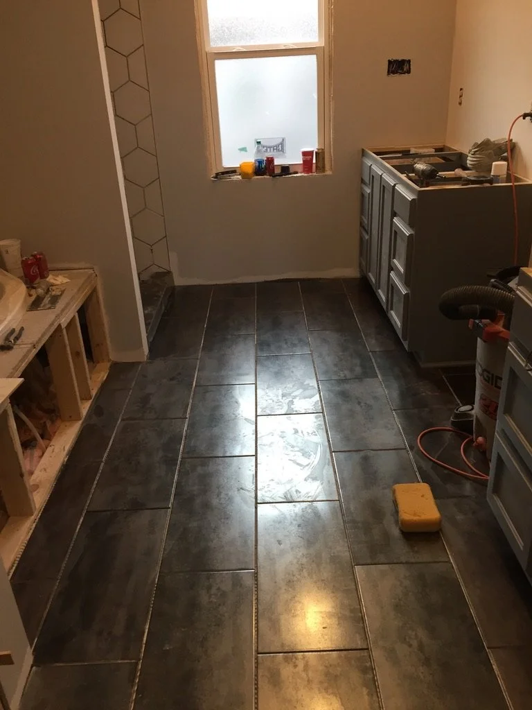 A room under renovation with dark tiled flooring, unfinished cabinetry, construction tools, and a window.