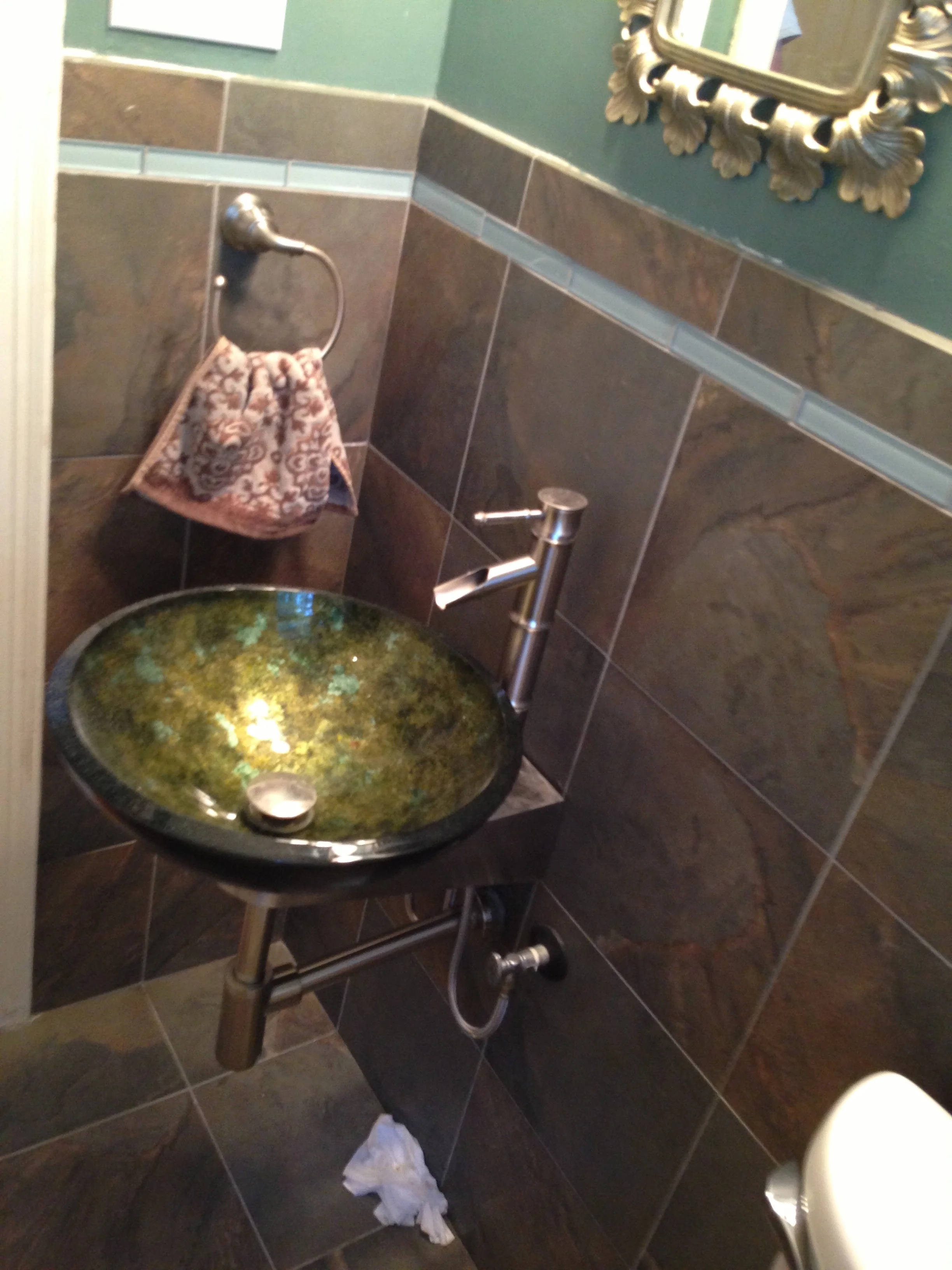A bathroom with a decorative vessel sink featuring green and black swirled pattern, a modern faucet, dark tiled walls, a mirror with an ornate frame, a towel ring with a floral towel, and paper on the dark tiled floor.