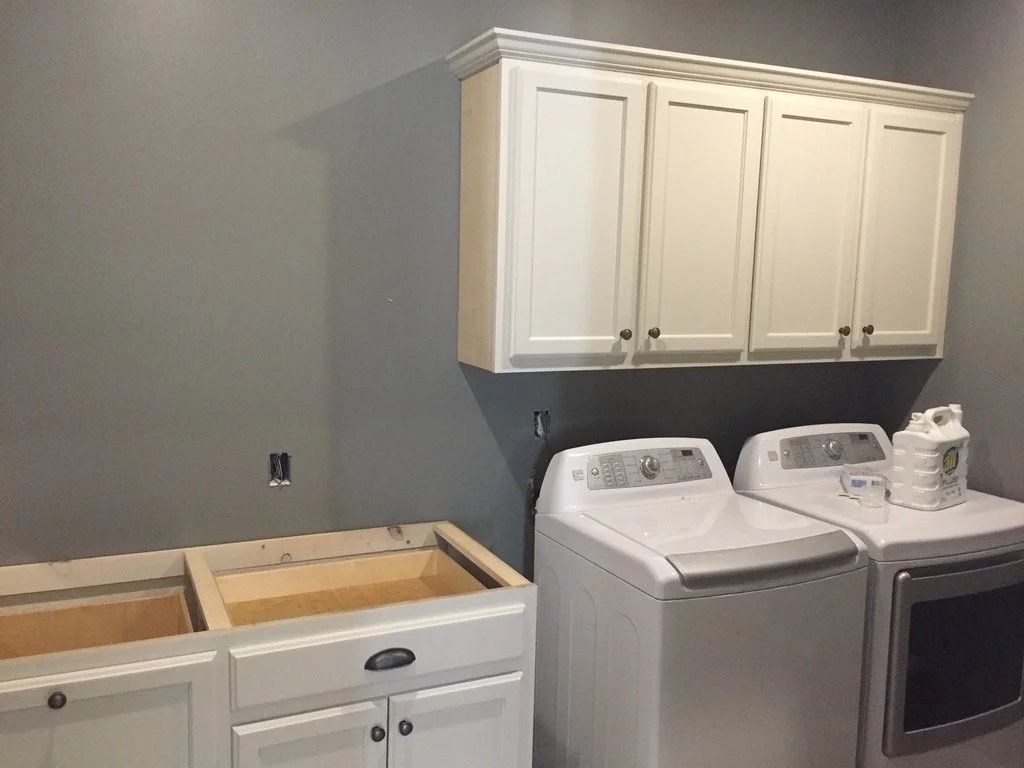 In-progress laundry room with white cabinets above washer and dryer, and partially installed lower cabinets awaiting countertop. Items include laundry detergent on the dryer.