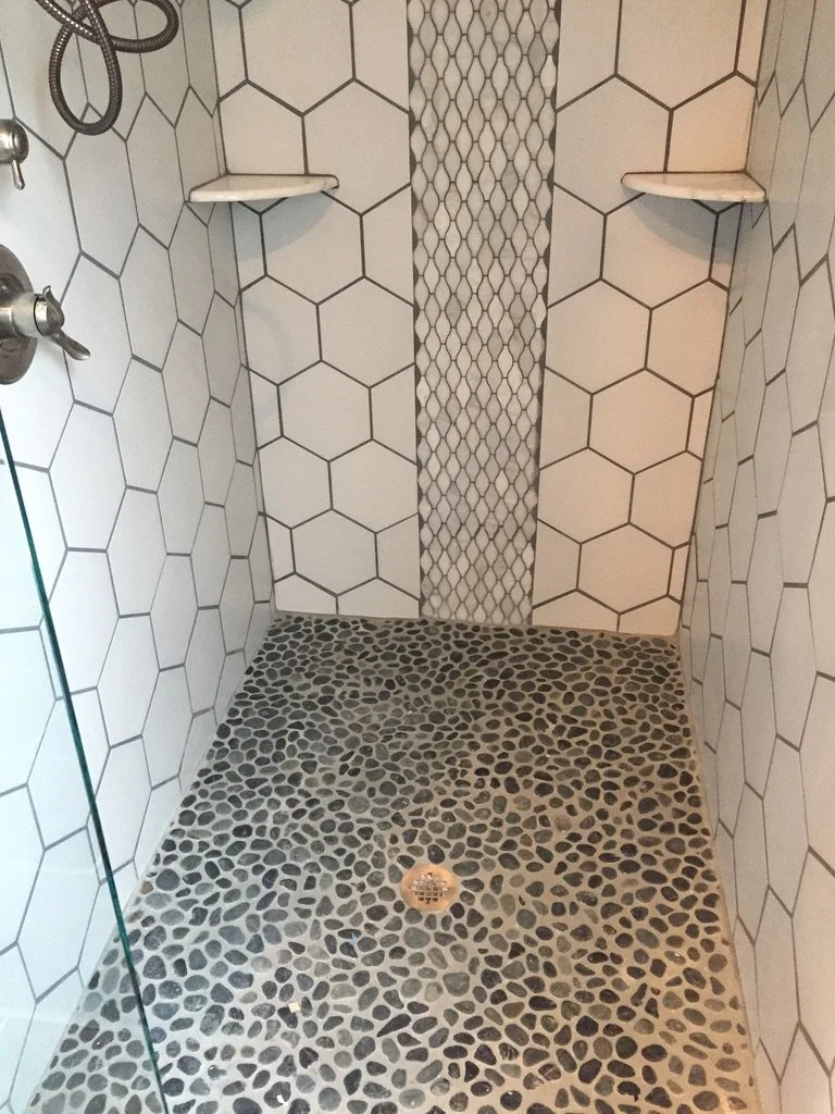 A tiled shower with white hexagonal wall tiles, a pebble stone floor, a central drain, a glass panel, and two corner shelves.