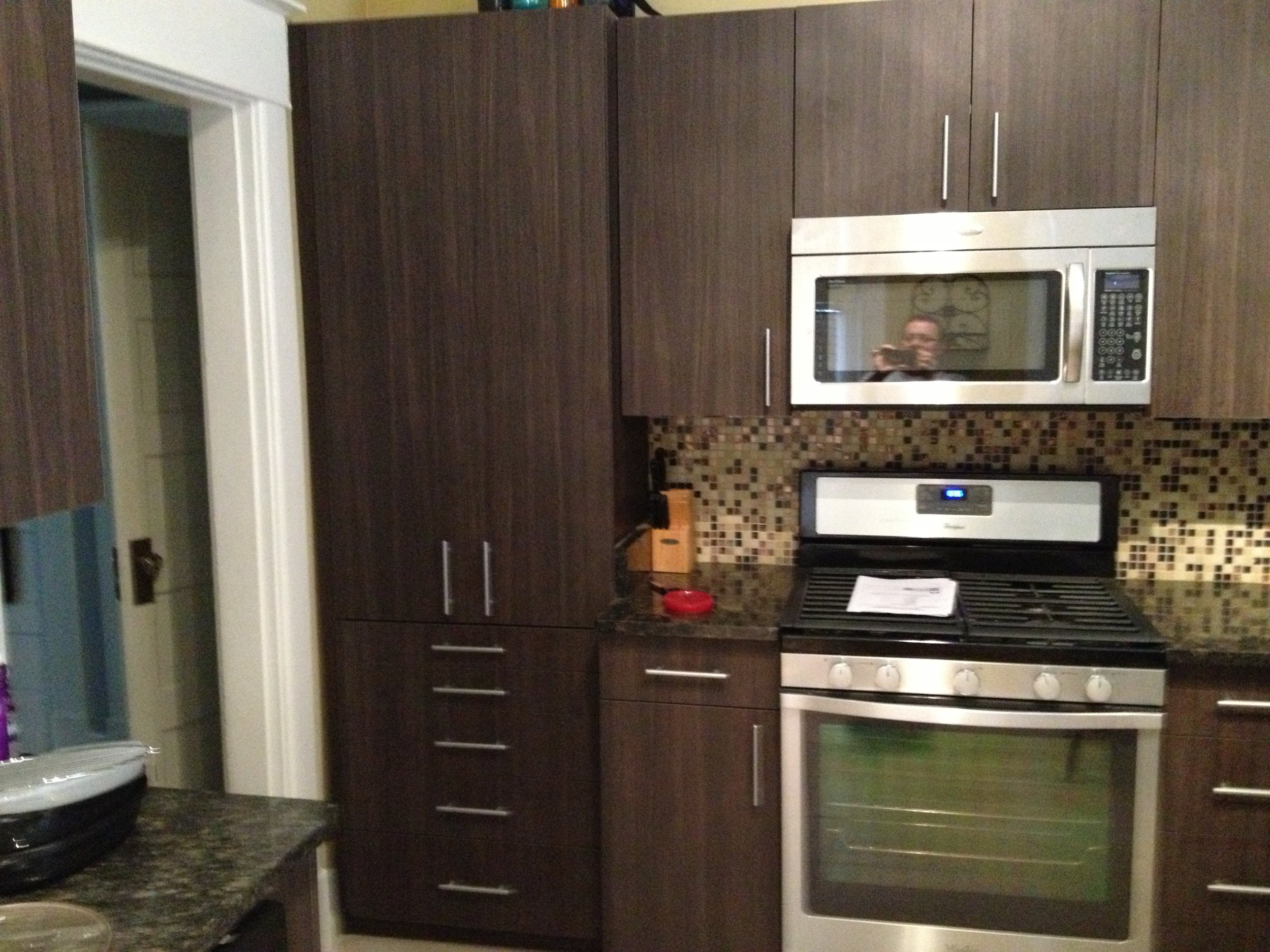 Kitchen with dark brown cabinets, a stainless steel microwave oven, a white oven, and a mosaic tile backsplash. There is a person reflected in the microwave door taking a photo.