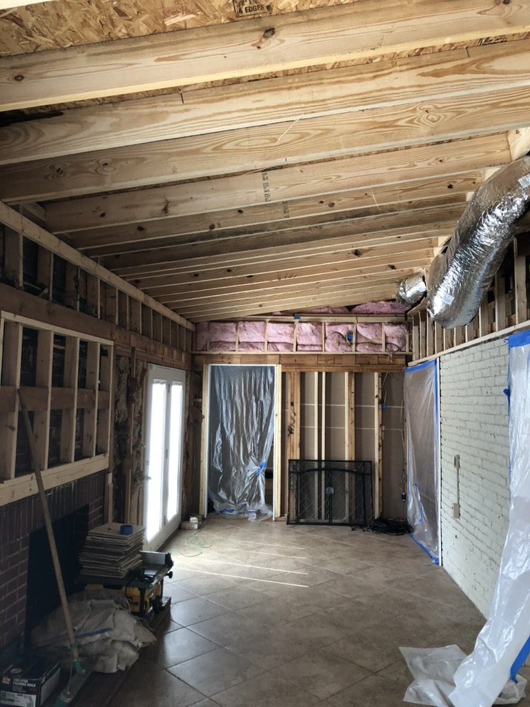 Interior of a room under construction, with wooden framing, insulation, and plastic sheeting covering some areas, and construction tools and materials on the floor.