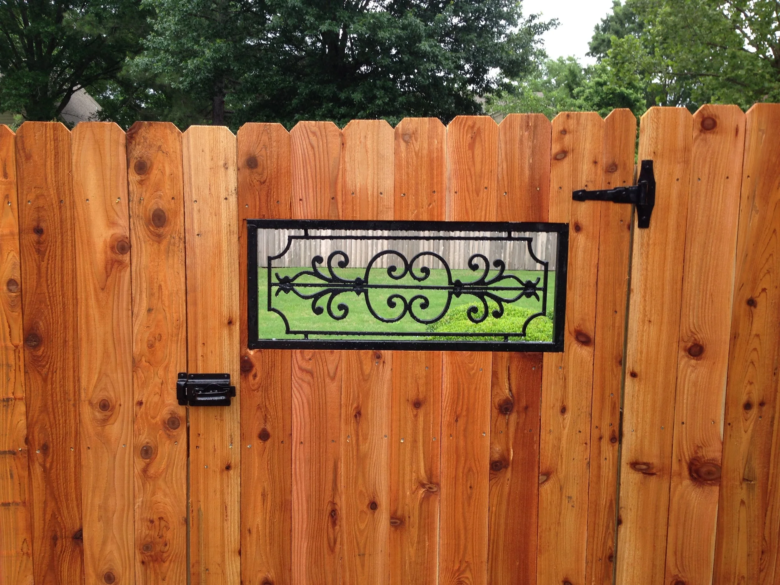 Wooden fence with a black iron gate, trees in the background, and a cloudy sky above.