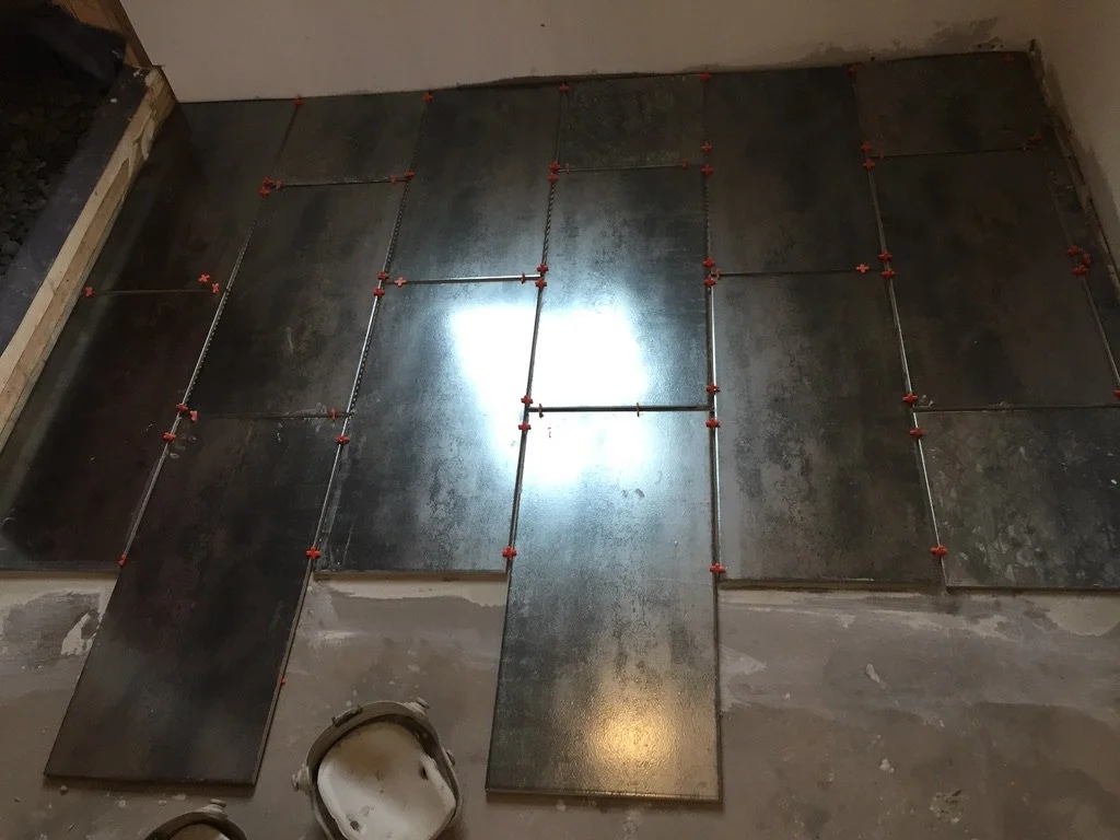 Underfloor heating pipes installed beneath dark ceramic tiles with red spacers in a room under construction.