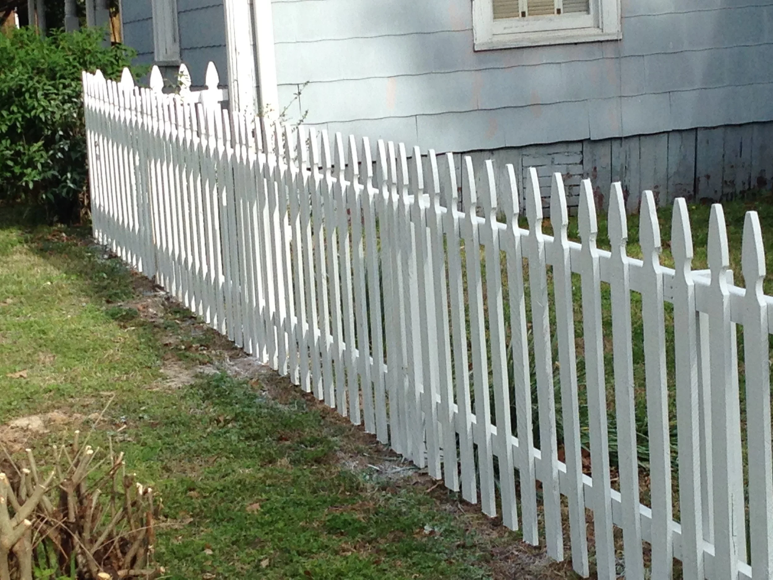 A white picket fence running along the side of a house with light blue siding and small windows. The fence is made of evenly spaced vertical boards with pointed tops. There is greenery, including grass and bushes, along the fence line.