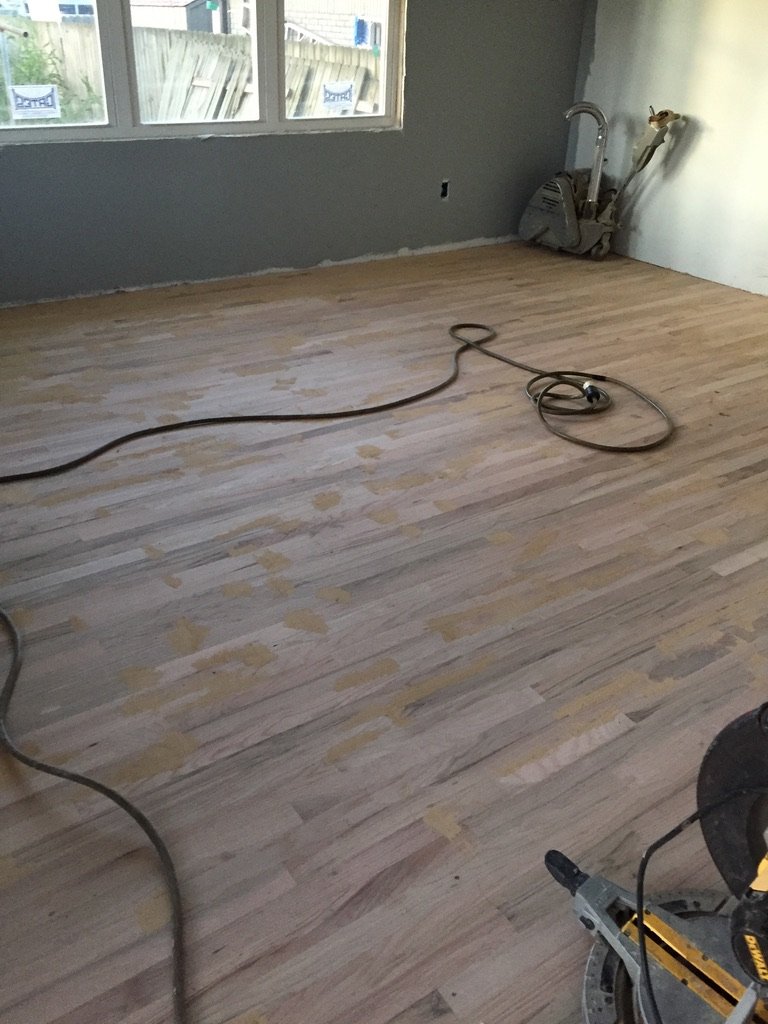Room with unfinished hardwood floor, construction tools and cord, gray wall, window, and white trim.