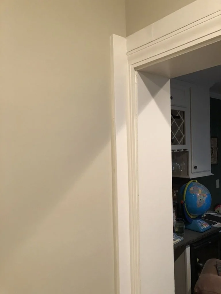 View of a beige wall with a white door frame, leading to a room with a dark countertop, a globe, and various items on the countertop.