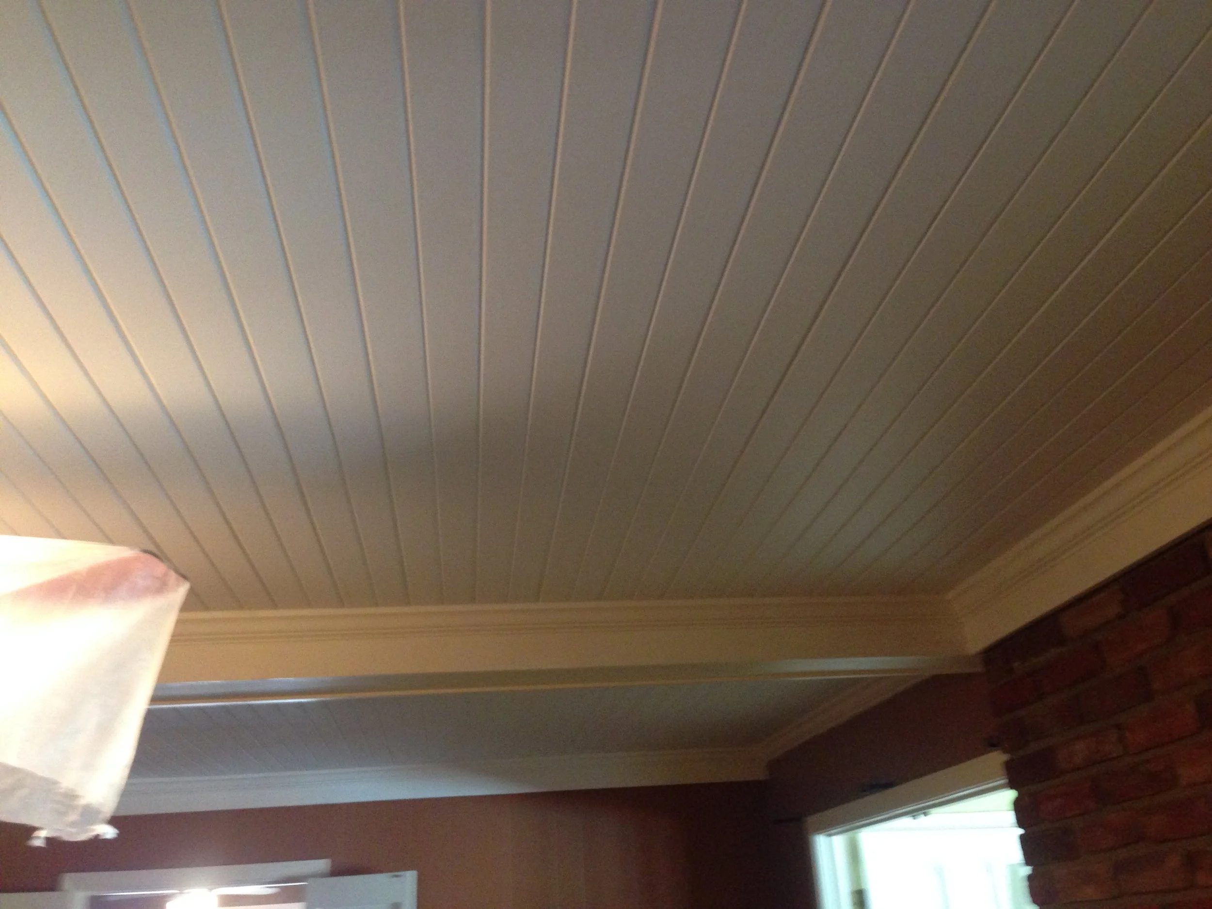 A ceiling with white wooden panels and trim, showing part of a curtain and window at the bottom right corner.