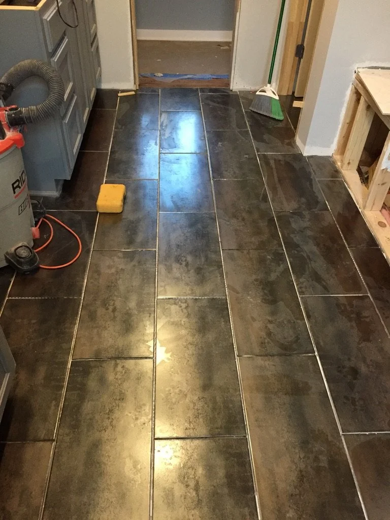 Newly installed dark ceramic tile flooring in a room, with construction tools and materials visible, including a sponge, a yellow block, a dustpan, and a vacuum hose.
