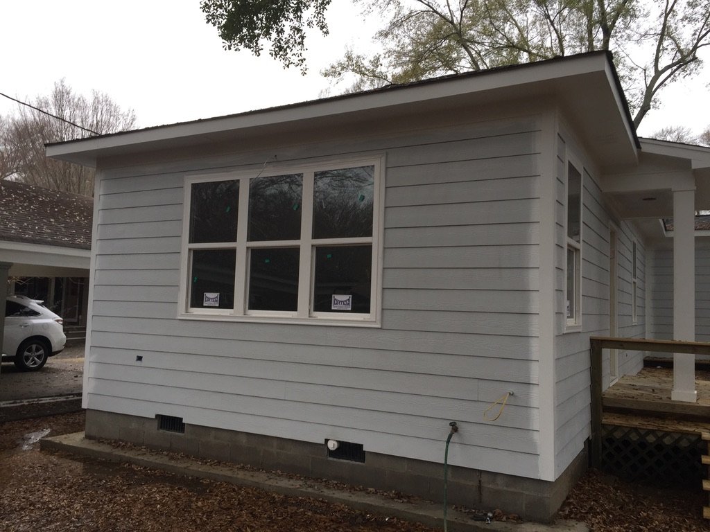 Newly constructed white house with horizontal siding, a large window, and a small porch area on the right. A car is parked in the driveway to the left.