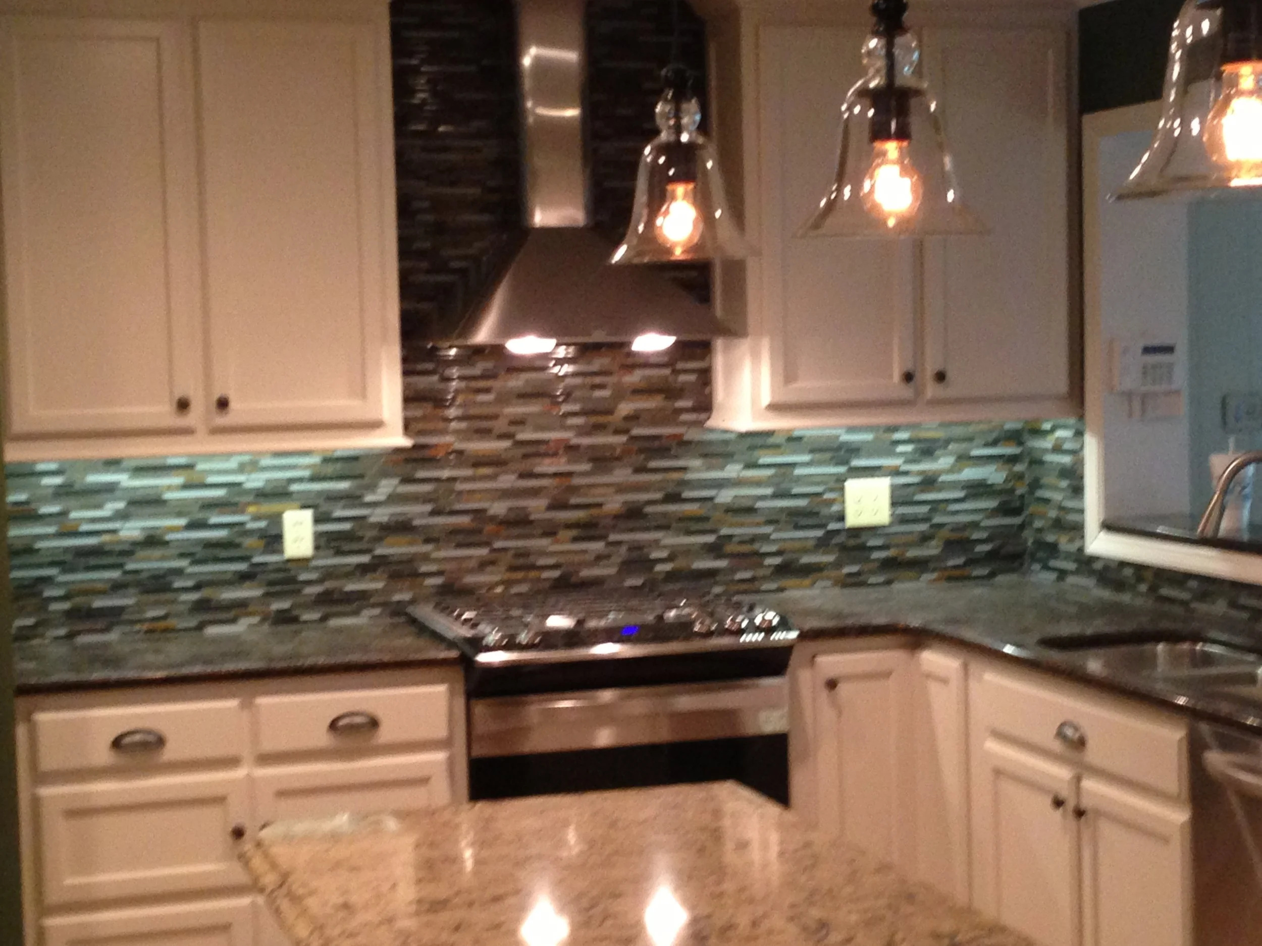 Kitchen with cream cabinets, a multicolored mosaic tile backsplash, a stainless steel range hood, and three pendant lights hanging above the counter.