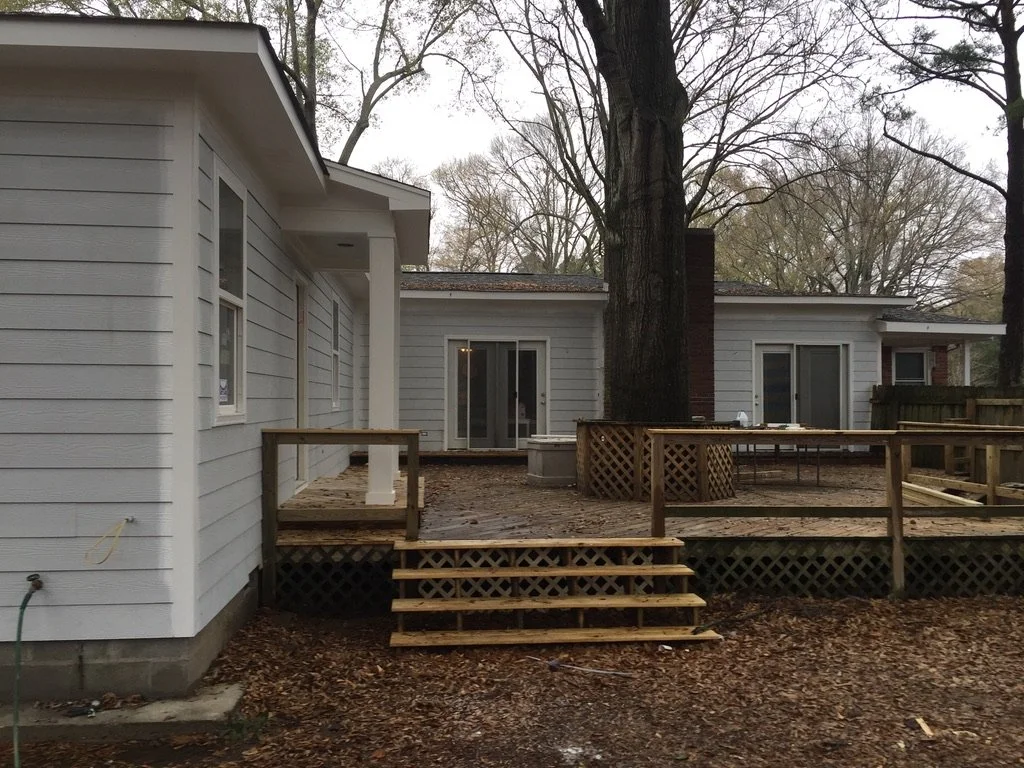 A backyard with a newly built wooden deck and steps, attached to a white house with sliding glass doors, surrounded by trees and fallen leaves.