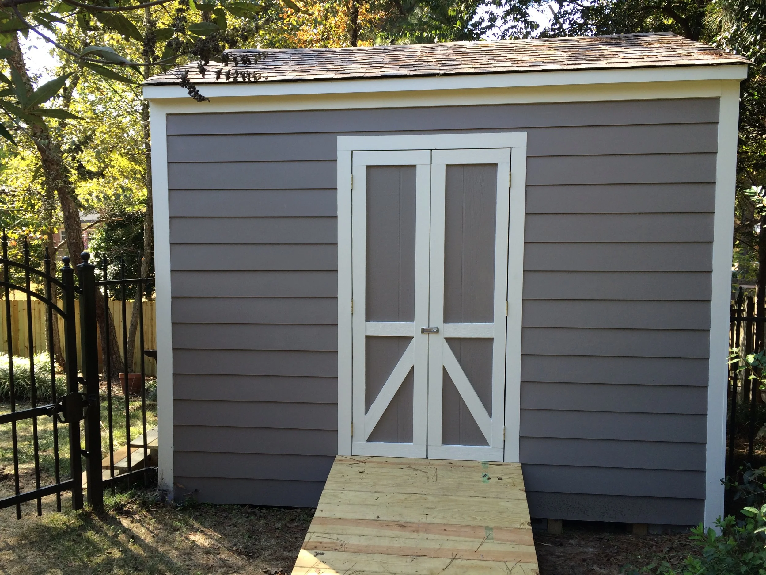A small gray shed with white trim and double doors, situated outdoors in a backyard surrounded by trees and a black metal fence. There is a wooden ramp leading up to the shed's door.