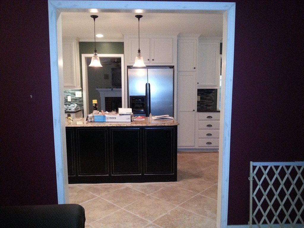 View of a kitchen seen through an open doorway, with white cabinets, black lower cabinets, and stainless steel appliances, including a refrigerator. There are two pendant lights hanging over a kitchen island.