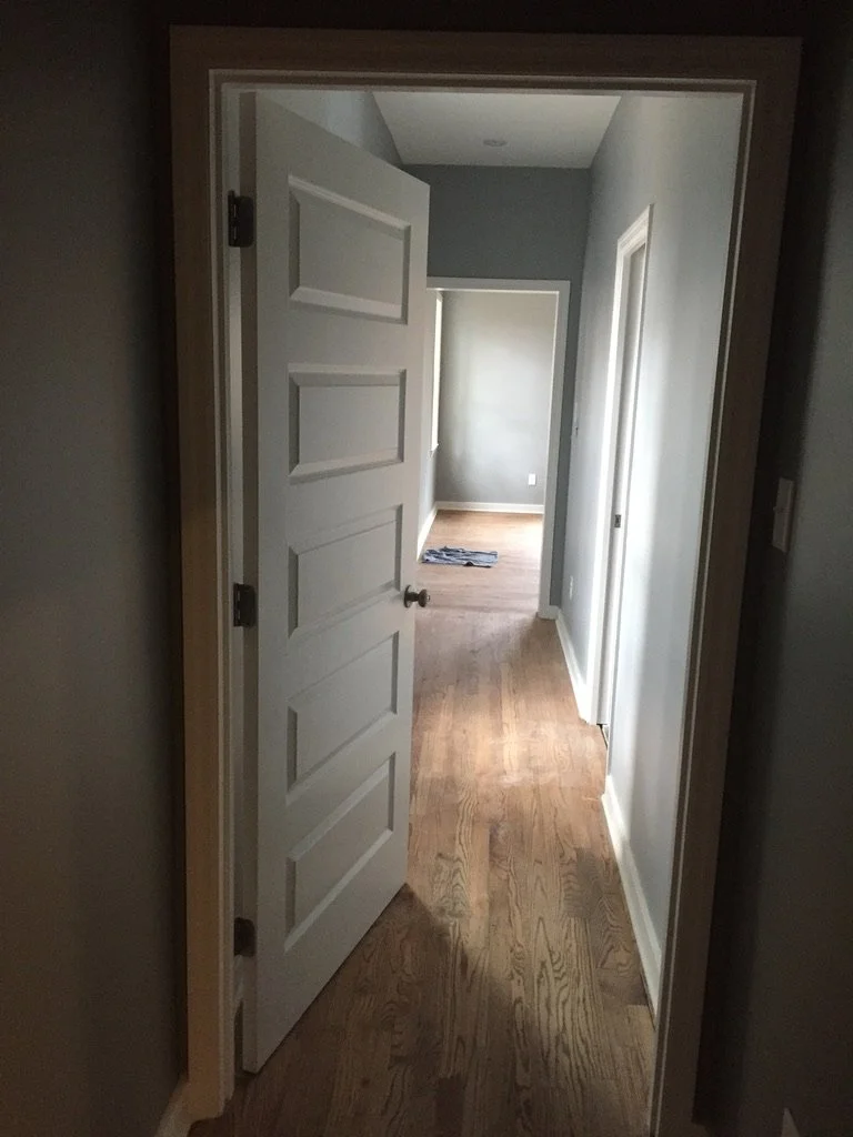 Interior view of a hallway with wooden flooring, gray walls, and white trim. An open white door reveals a room with a window and a gray item on the floor.