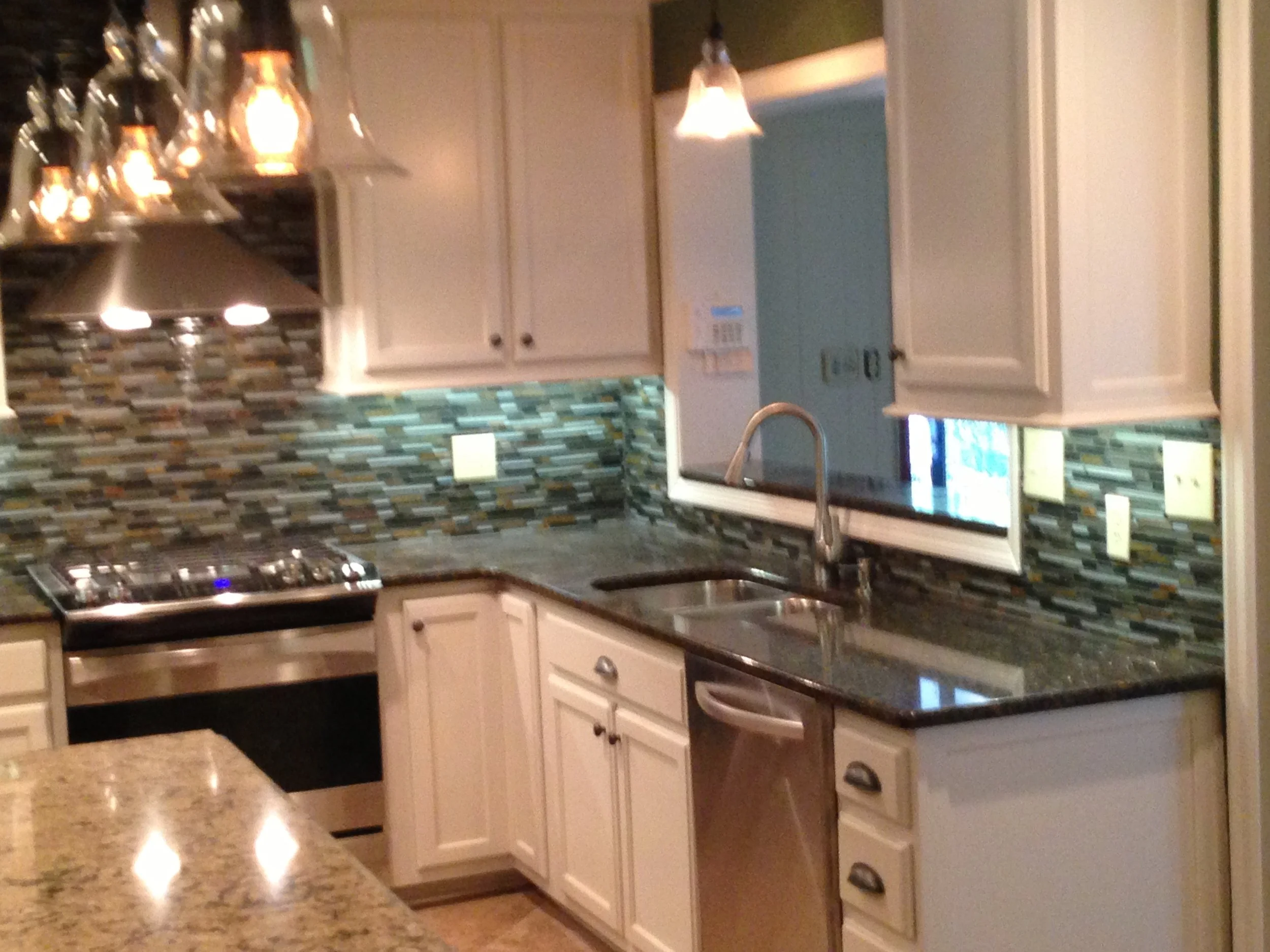A kitchen with beige cabinets, granite countertops, a mosaic tile backsplash, a stainless steel stove, a double sink, and pendant lights hanging above.
