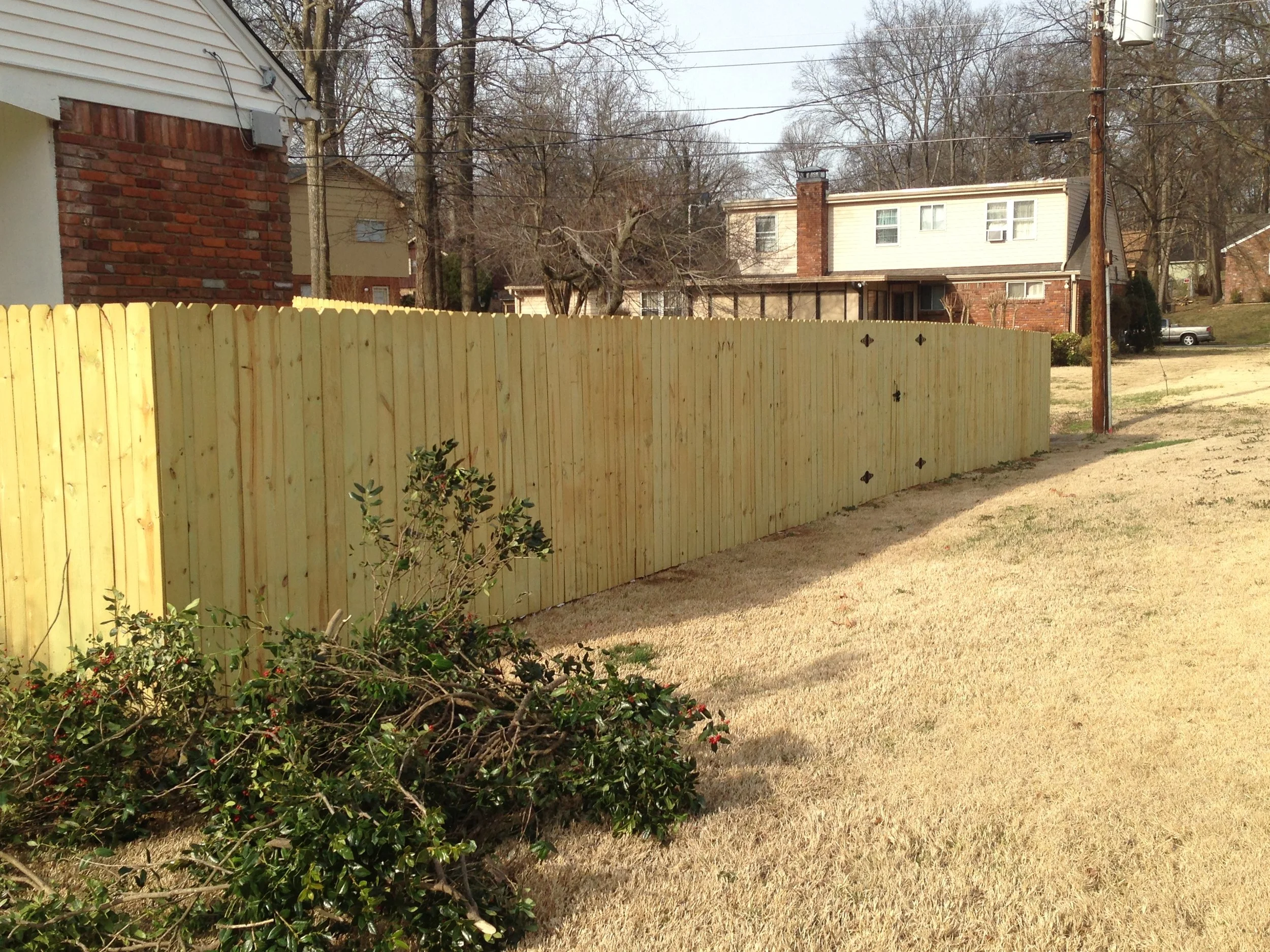 A view of a suburban backyard with a tall wooden privacy fence, leafless trees, a two-story house with a brick chimney, some electrical wires and poles, and a parked car in the driveway.