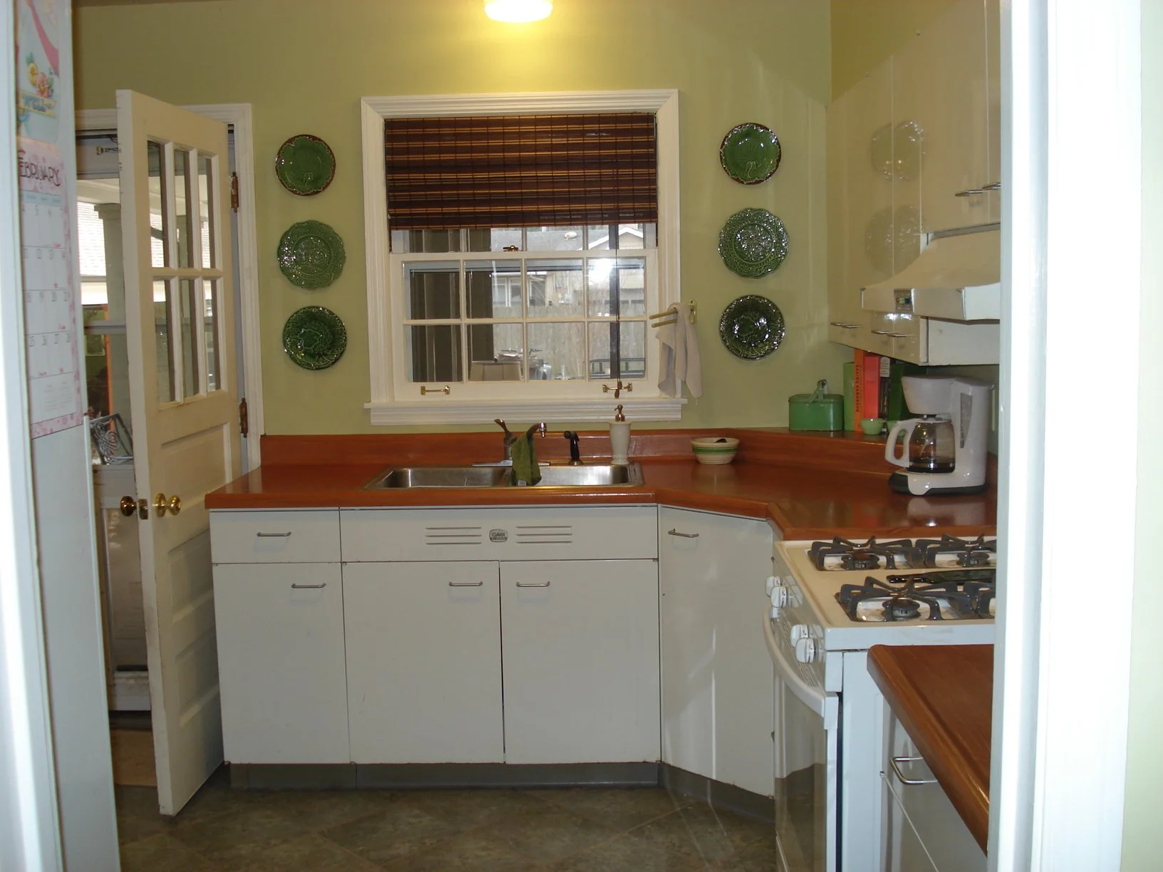 A small kitchen with white lower cabinets, wooden countertops, and green decorative plates on the mustard-colored wall. There is a window above the sink with blinds, a coffee maker, a green container, and books on the right side. A white stove is als