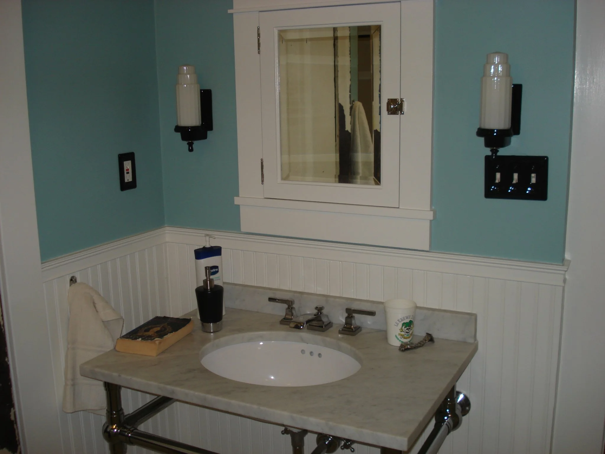 Bathroom sink with mirror cabinet above, soap dispenser, and various toiletries on the marble countertop, with a wall-mounted hairdryer and electrical outlets nearby.