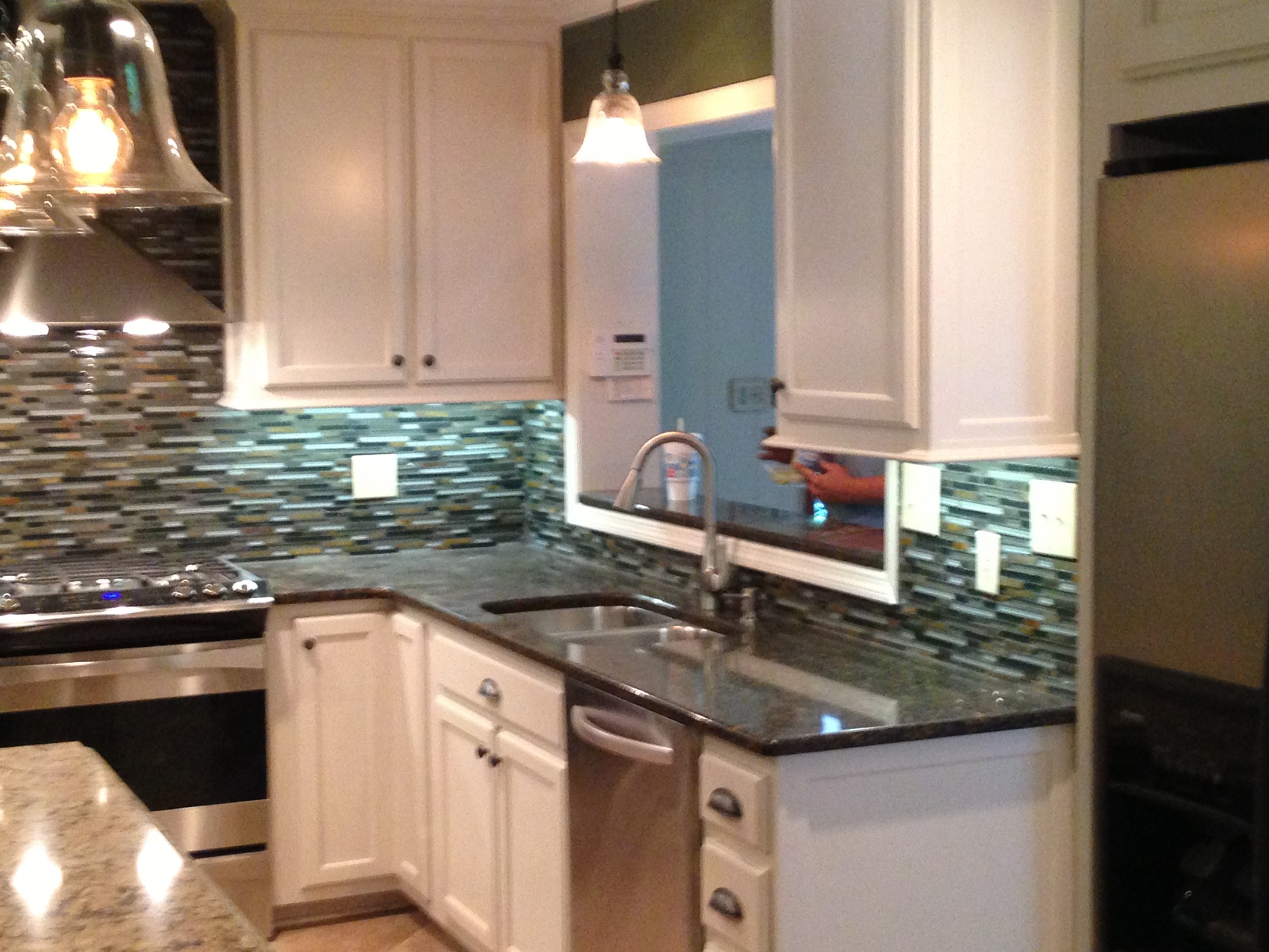 A kitchen with white cabinets, a granite countertop, a colorful mosaic tile backsplash, a stainless steel stove, and a sink. There is a person in the background holding a phone.