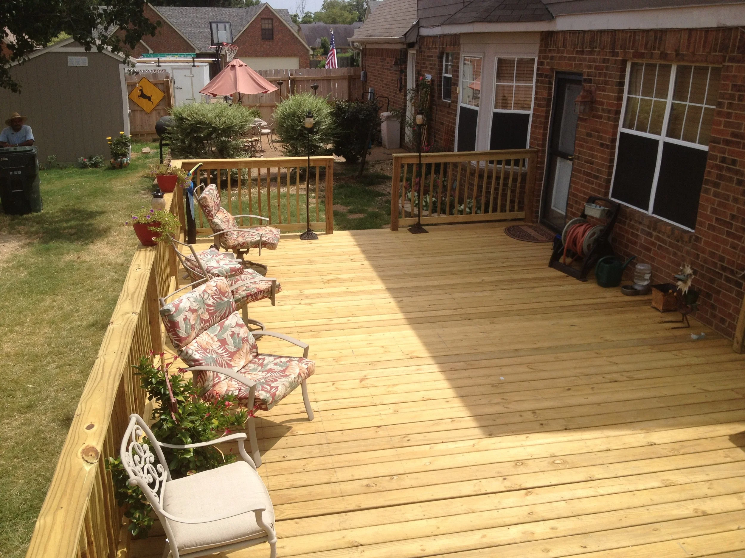 A backyard with a wooden deck and a grassy lawn. There are chairs with floral cushions on the deck, potted plants, and a railing. The yard has bushes, a table with an umbrella, and a basketball hoop. A person wearing a straw hat is near a trash bin, 