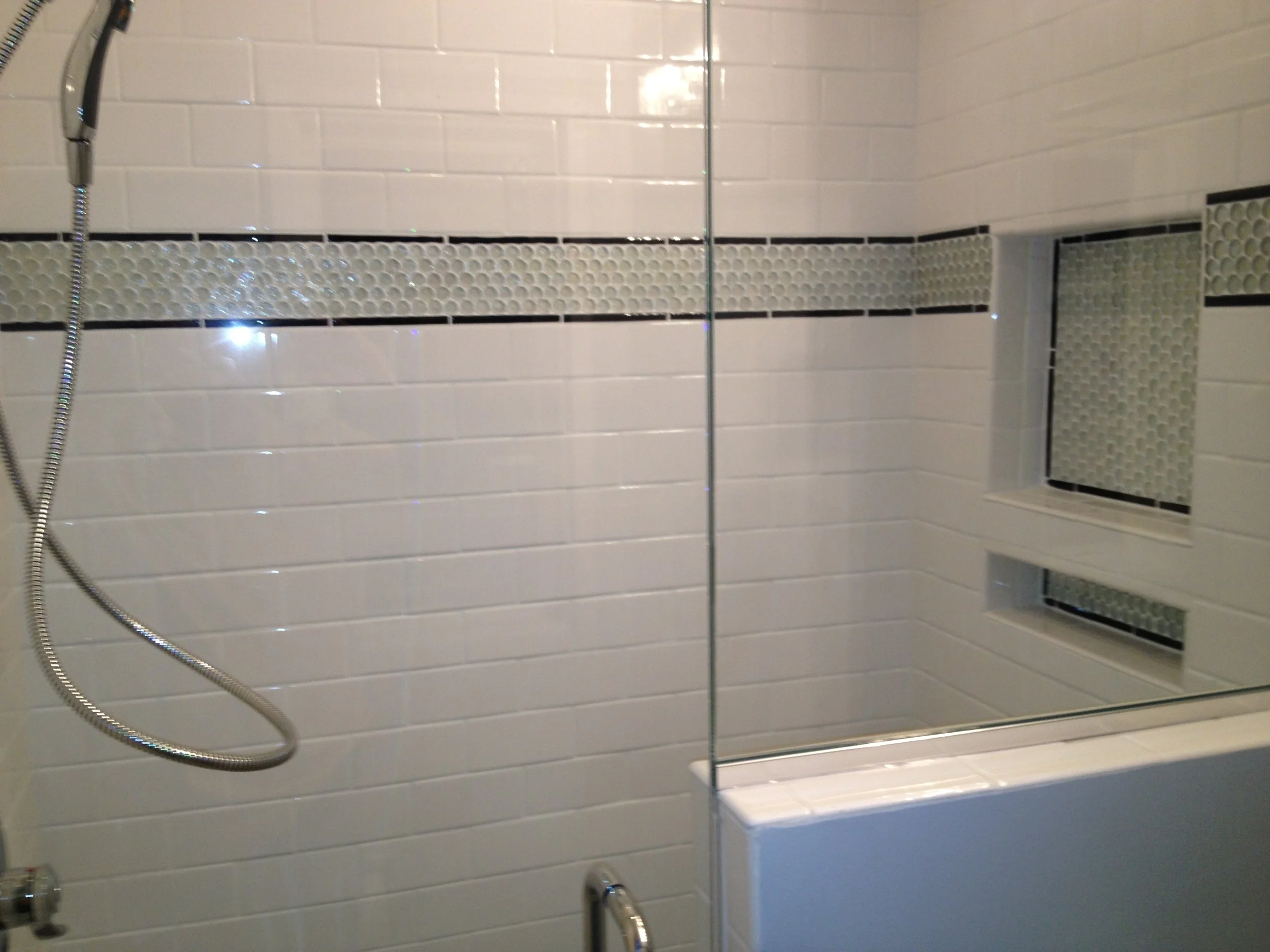 Shower area with cream-colored subway tiles, a horizontal strip of textured circular tiles, a wall mirror, a handheld showerhead with a flexible hose, a window with a screen, and a tiled shower base.