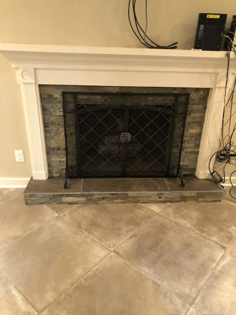 A fireplace with a white mantel, a brick interior, and a black metal fireguard. There are electrical wires and devices on top of the mantel and on the side.