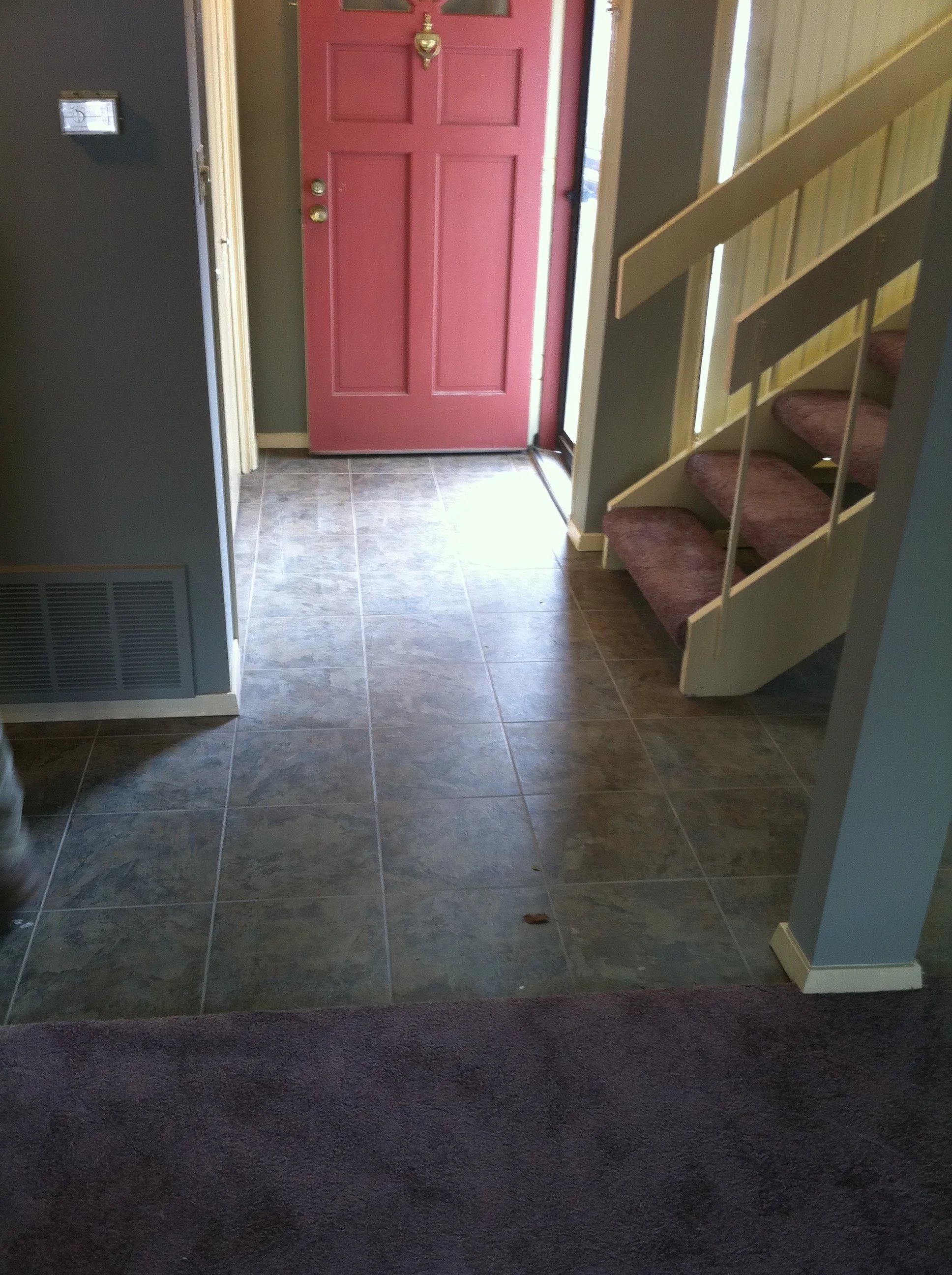 This image shows a hallway with a pink front door, a staircase with red carpeted steps, and a tiled floor. The door has a brass door knocker and door knob. The walls are painted in neutral colors, and a door frame is visible on the left side.