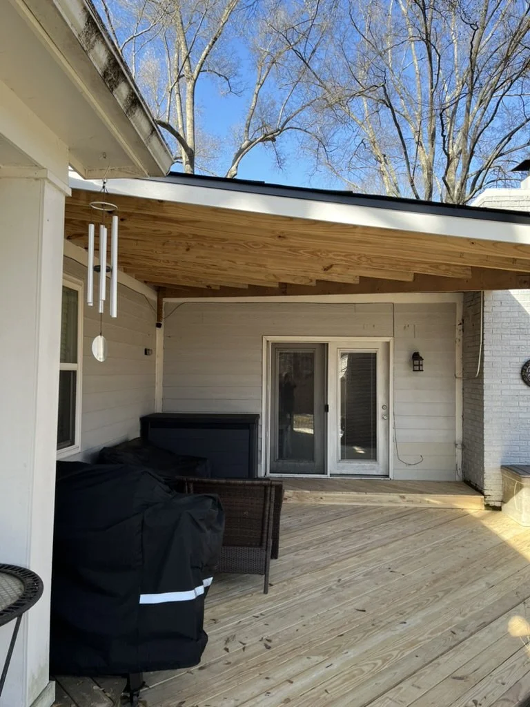 Back porch with wooden deck, patio furniture, and a grill covered with a black cover. There are sliding glass doors leading into the house and a wind chime hanging from the porch roof. Bare trees are visible in the background.