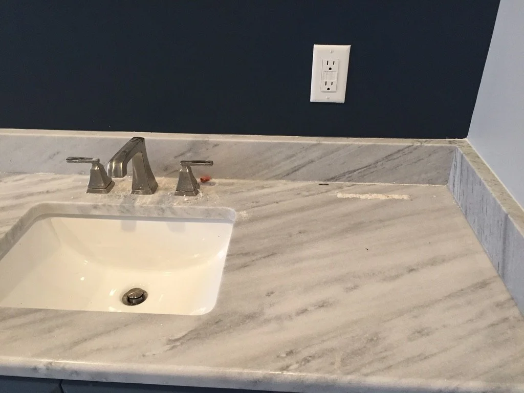 Bathroom countertop with a marble surface, a sink, a chrome faucet with two handles, a wall outlet above the countertop, and dark blue and light gray walls.
