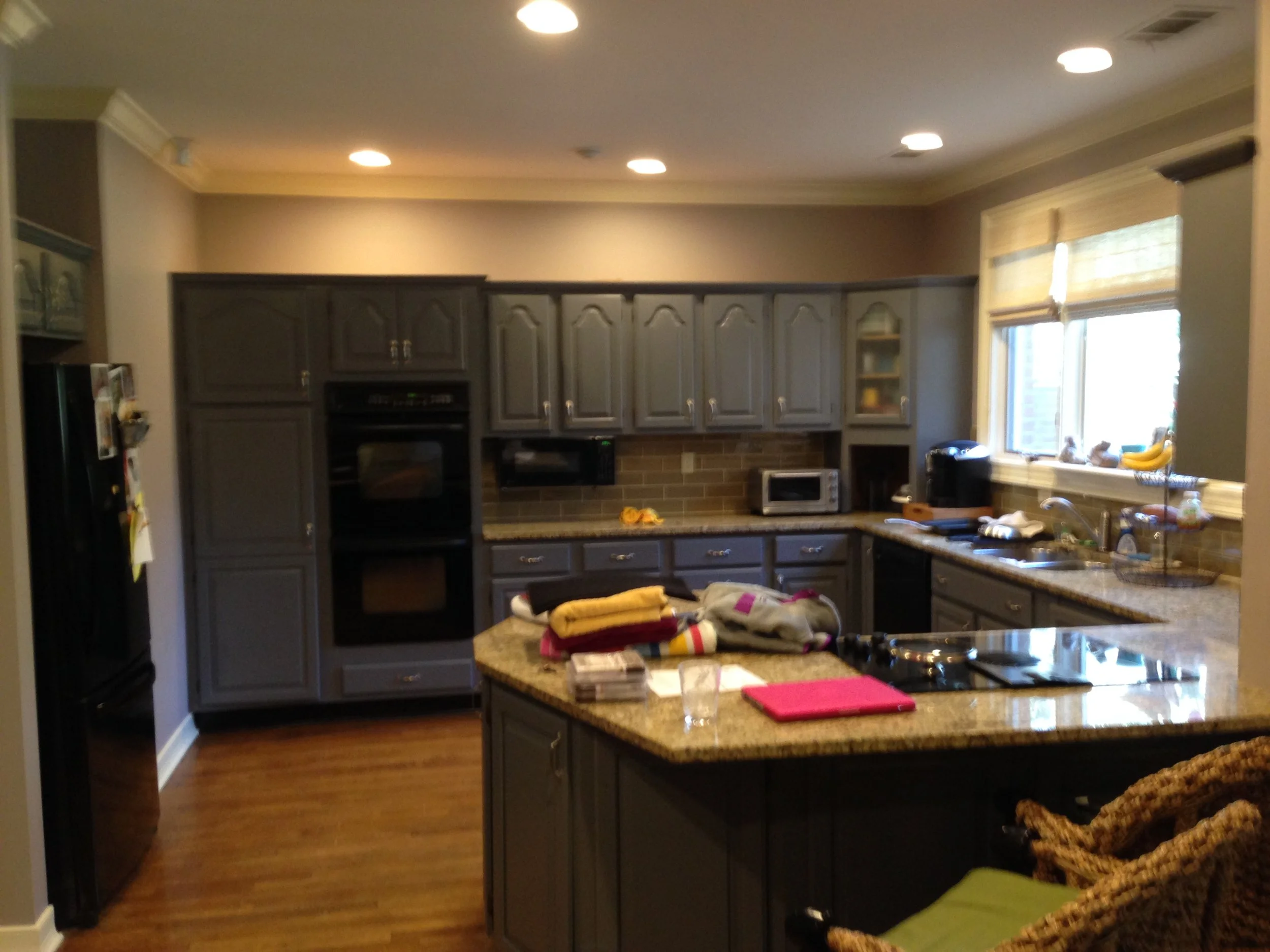 A kitchen with upper gray cabinets, a granite island countertop, and a window with blinds. Items on the counter include a microwave, a toaster, fruit, and various small objects.
