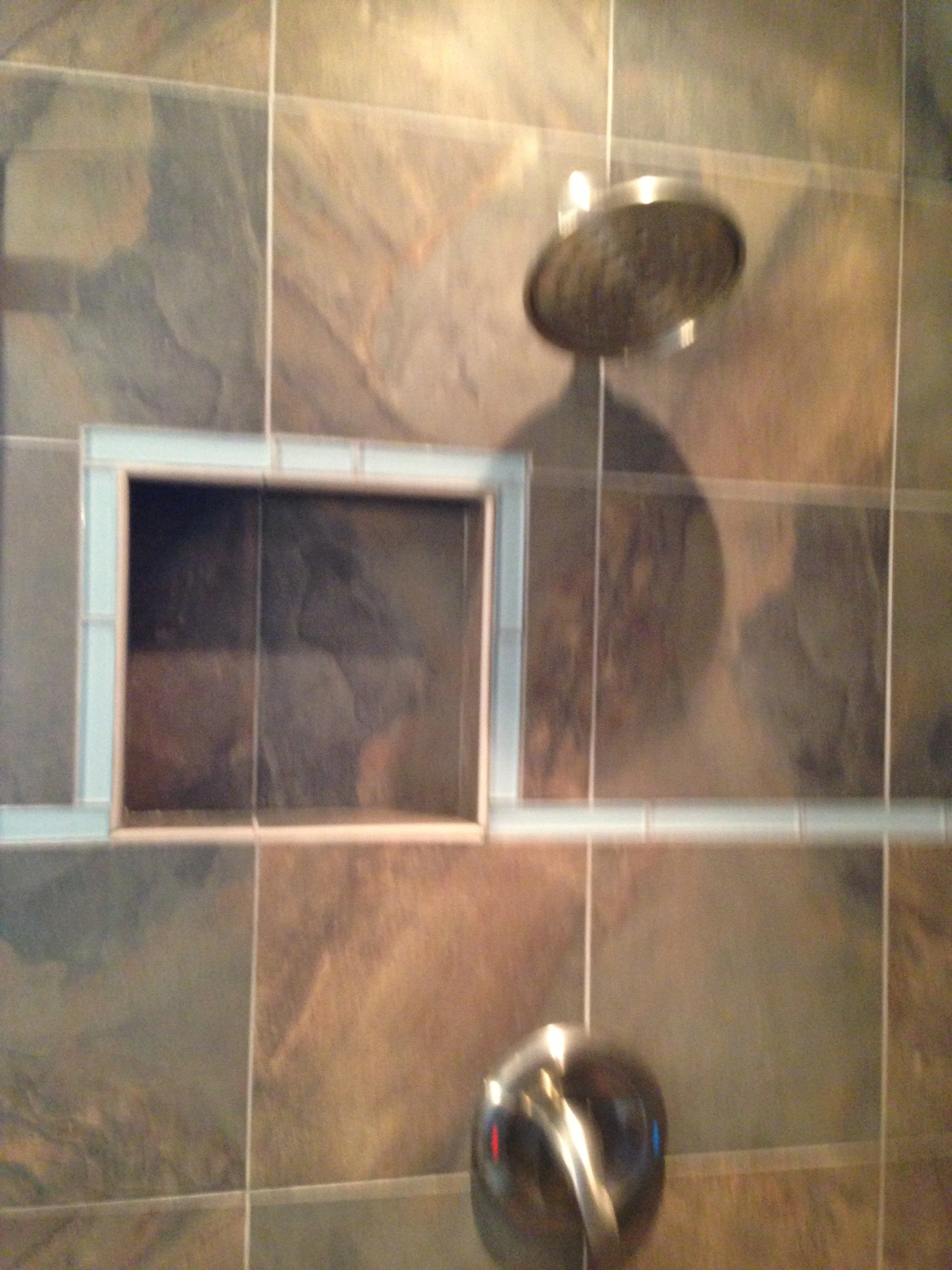 A bathroom with dark brown and tan tiled walls, showing a black flush button for a toilet and part of a flush tank installed in a tiled wall.