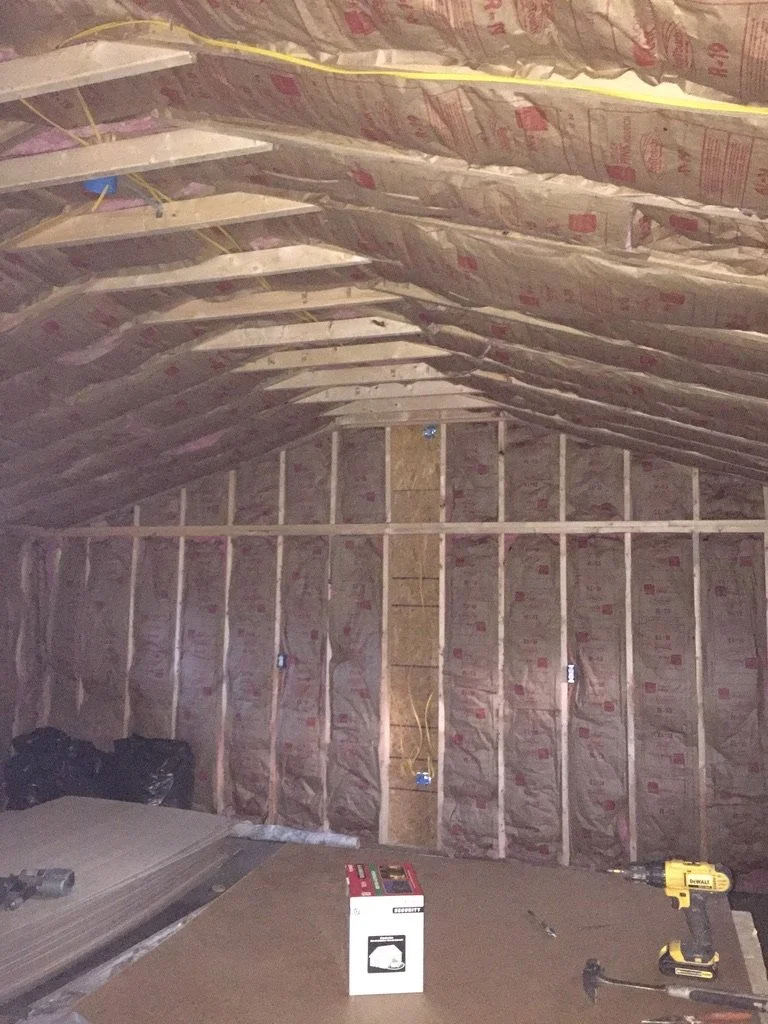 Construction of an unfinished room with exposed insulation, electrical wiring, and framing. A yellow drill, a box, and other tools are on a work surface.