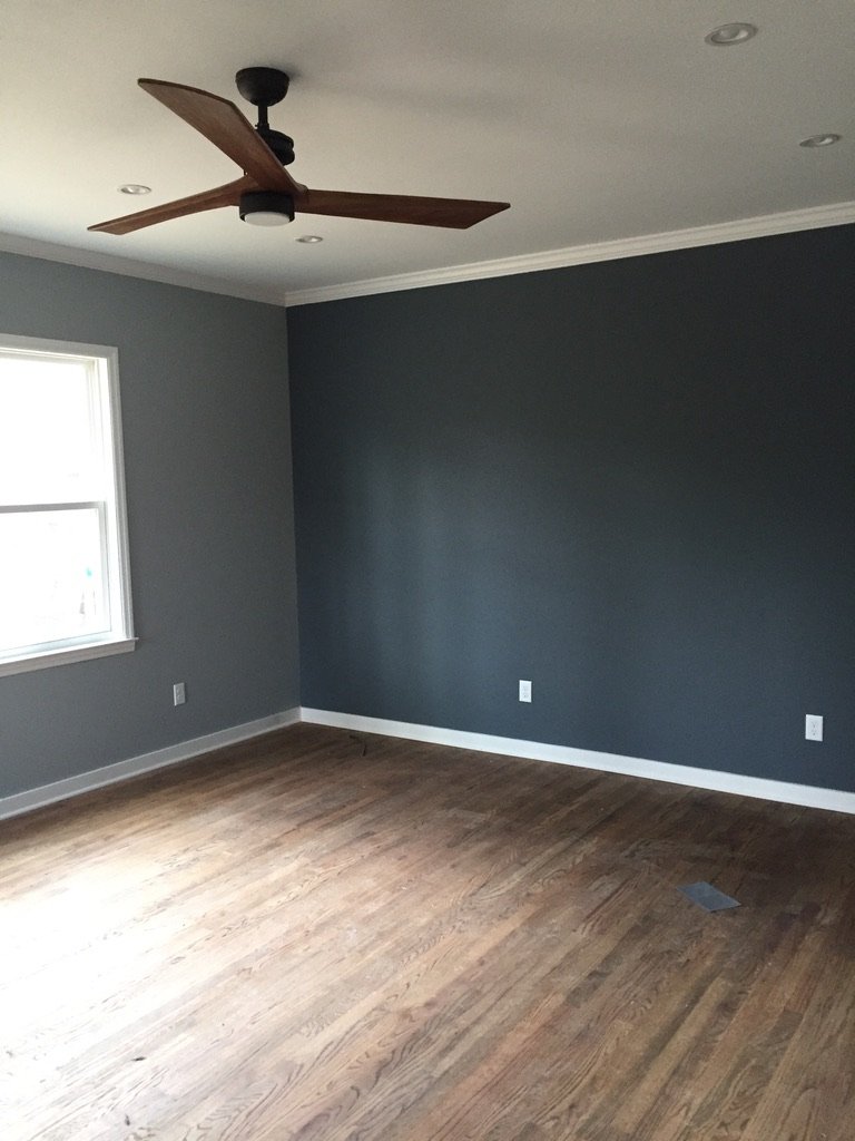 Empty living room with dark gray walls, hardwood floor, a ceiling fan, window, and white baseboards.