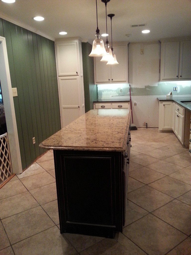 Kitchen with white cabinets, green accent wall, granite countertop island, and pendant lights overhead. Some cabinets appear to be unfinished or under renovation.