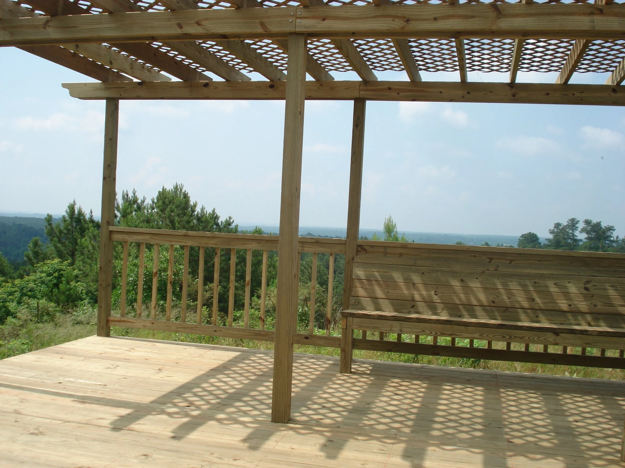 Wooden deck under construction with partial railing, surrounded by trees and a distant view of the landscape under a partly cloudy sky.