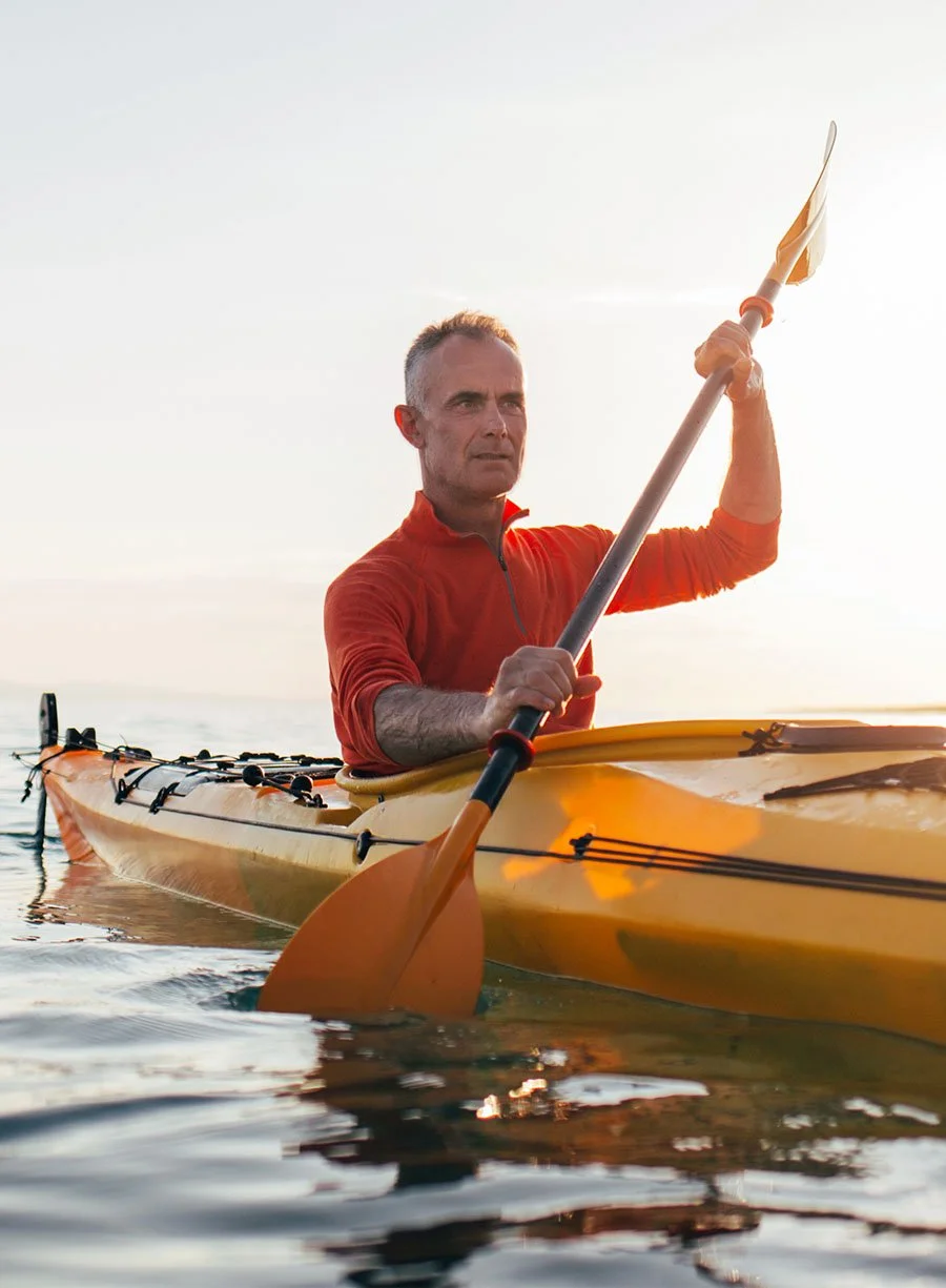 A middle-aged man paddling a yellow kayak on the water during sunset, wearing a red long-sleeve top.
