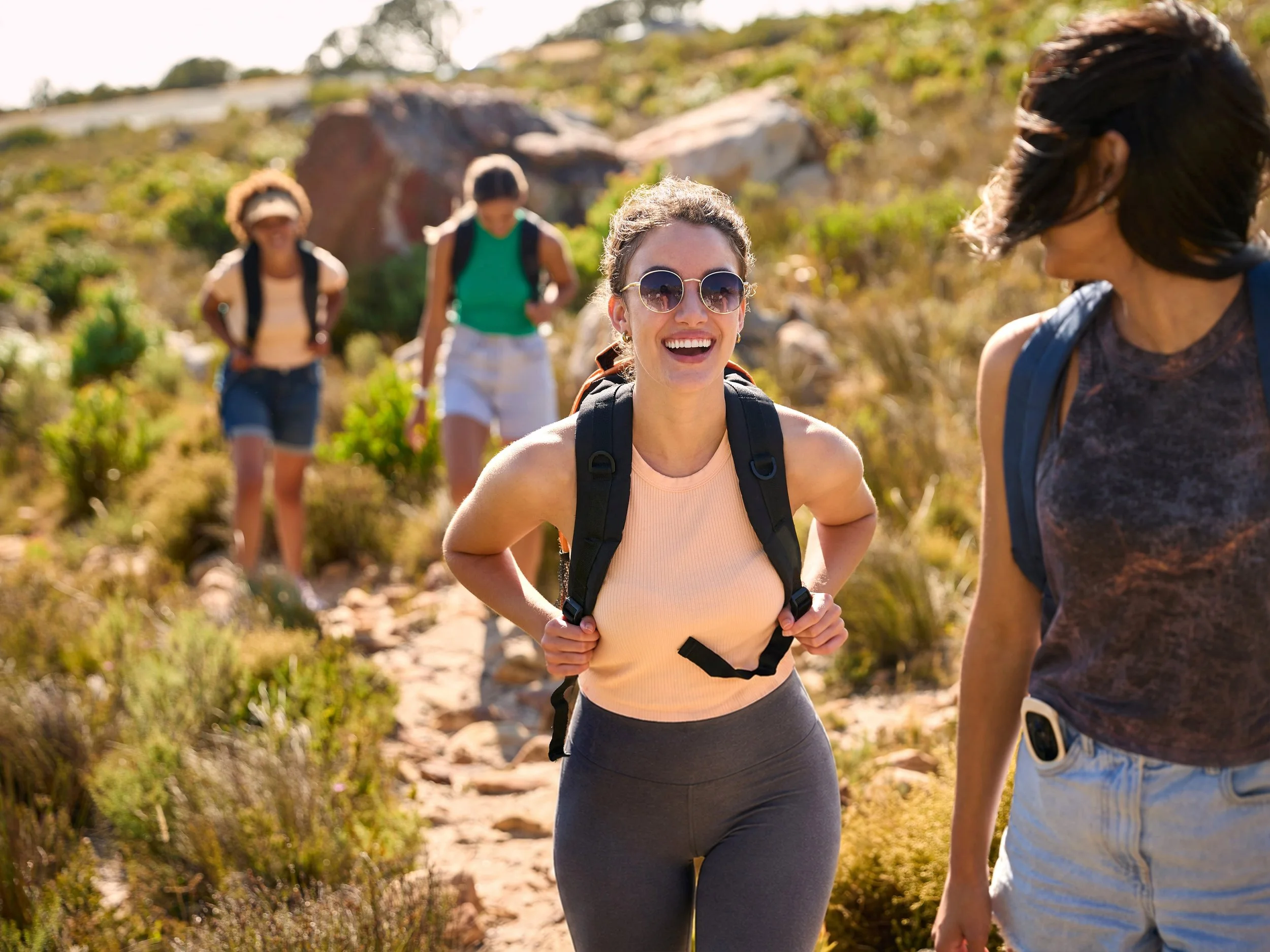Four women hiking on a sunny trail, smiling and chatting, wearing backpacks and athletic clothing.
