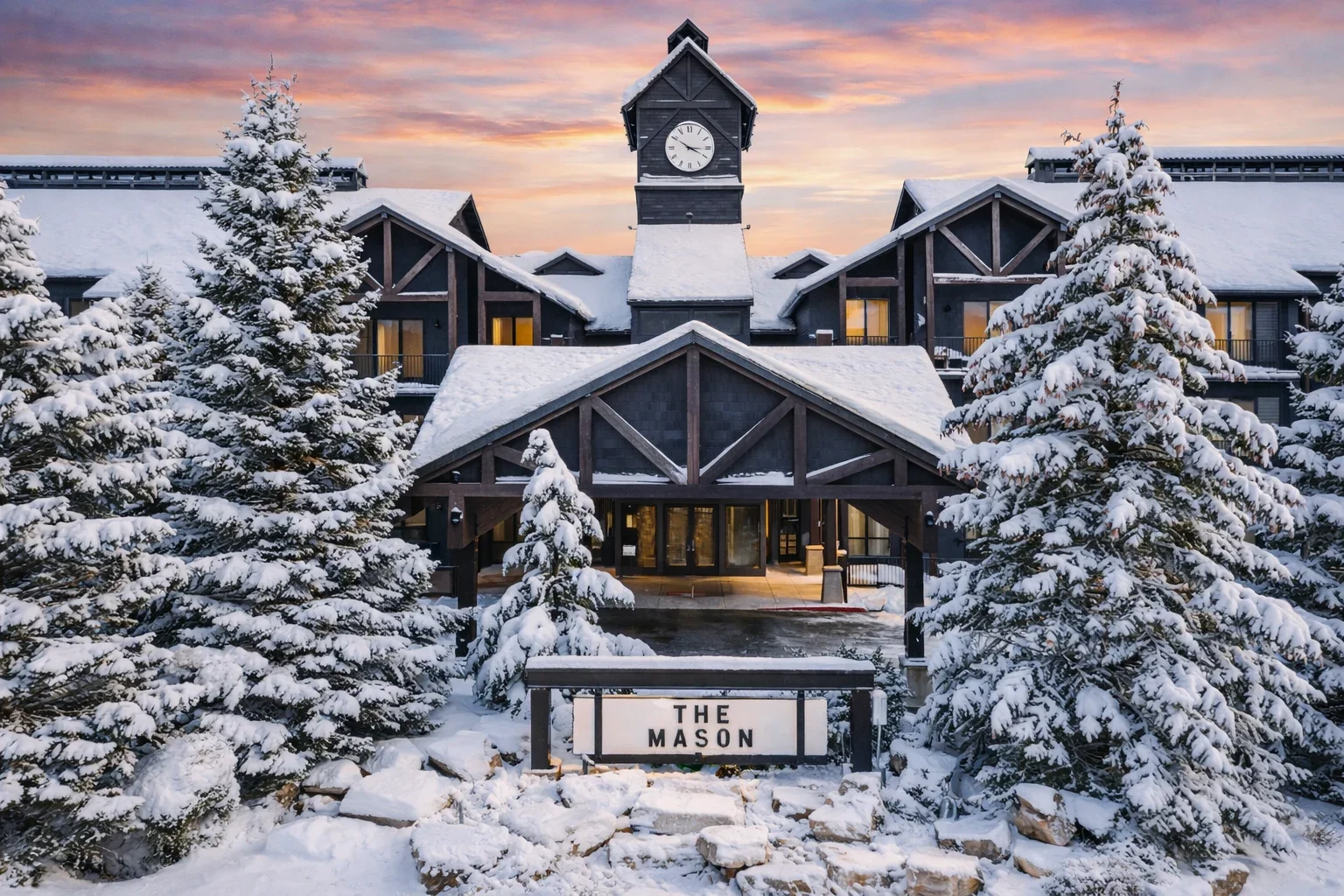 Snow-covered lodge named The Mason with snow-laden trees in front, a clock tower on top, and a colorful sky at sunset.