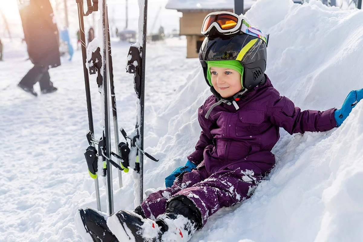 A young child dressed in purple snow gear and a black helmet with goggles sitting in the snow beside skis in a winter setting.