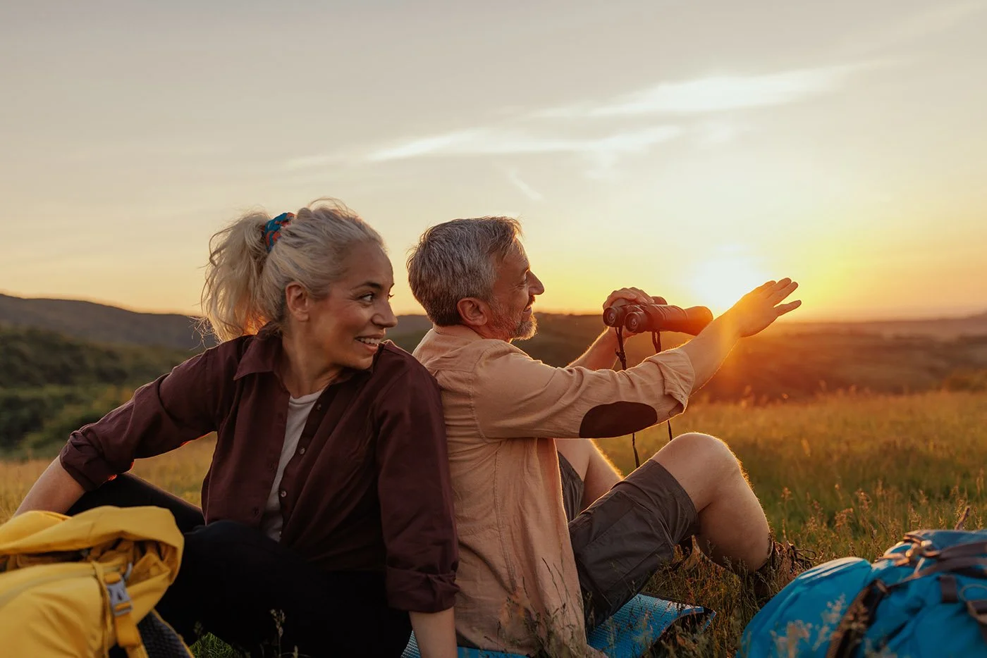 A woman and a man sitting on grass during sunset, with the man holding binoculars and pointing towards the horizon.