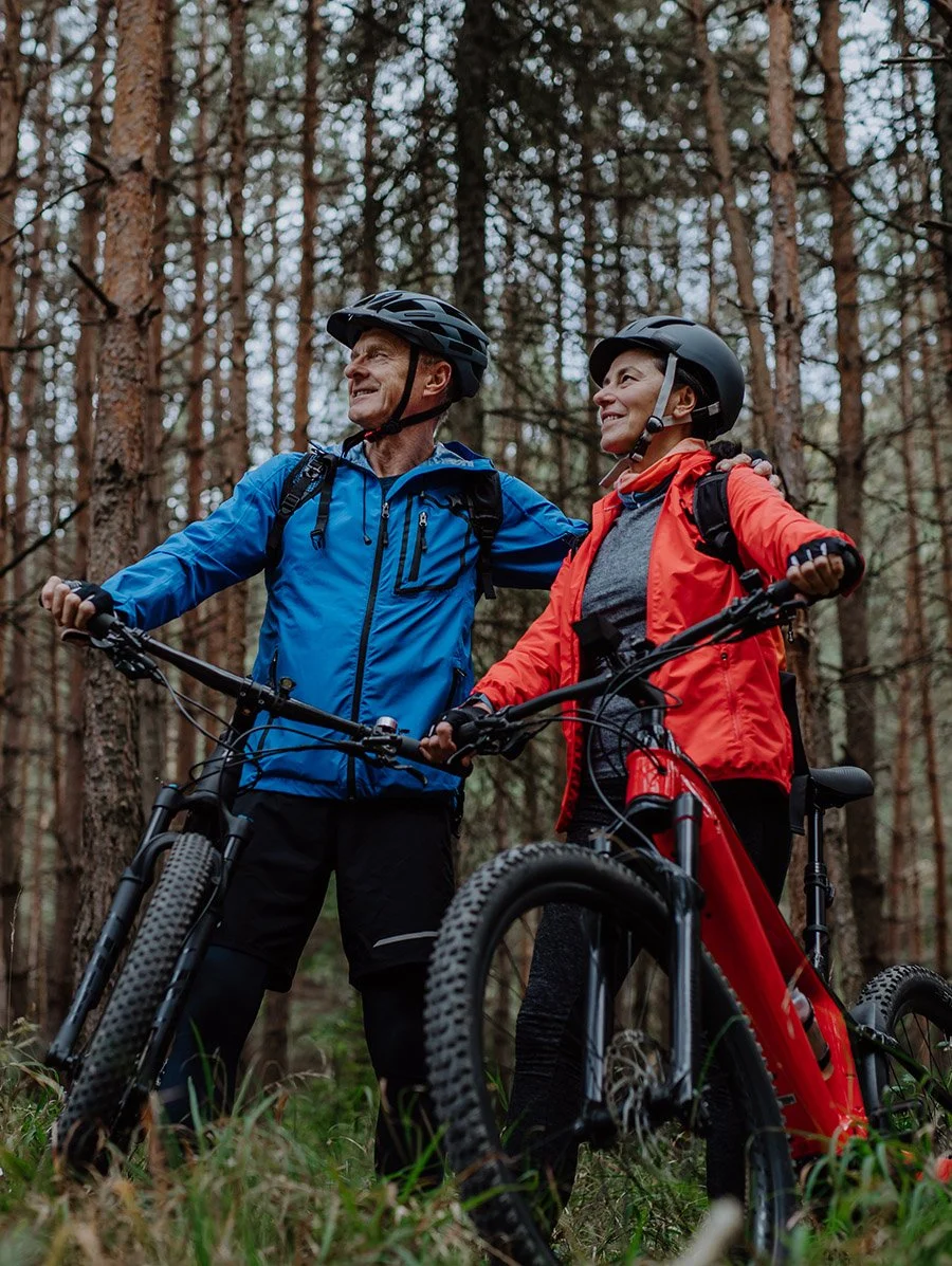 Two older adults, a man and a woman, in outdoor gear standing with their mountain bikes in a wooded forest, smiling and looking at each other.