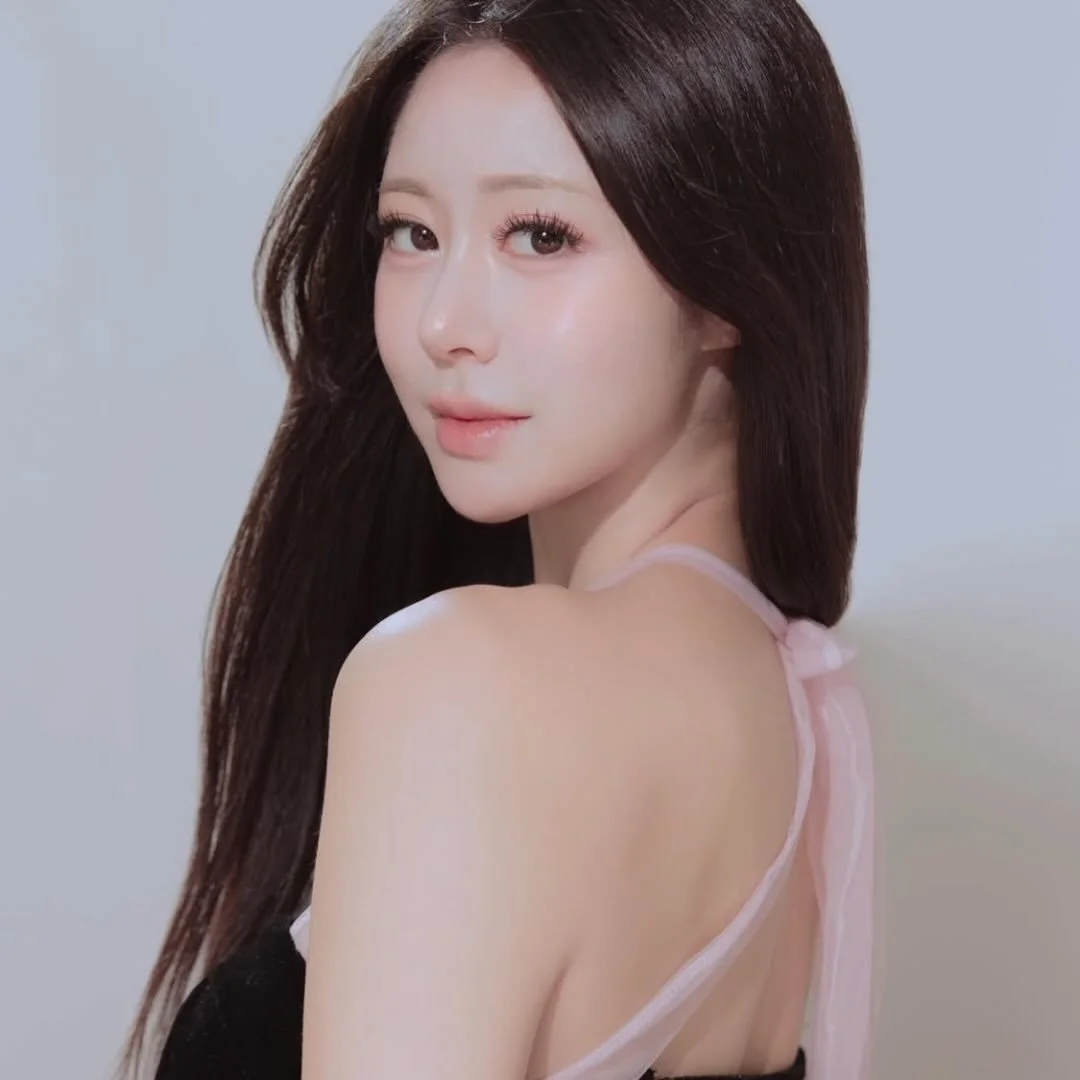 A young woman with long brown hair, wearing a black top with pink accents, looks over her shoulder with a slight smile.